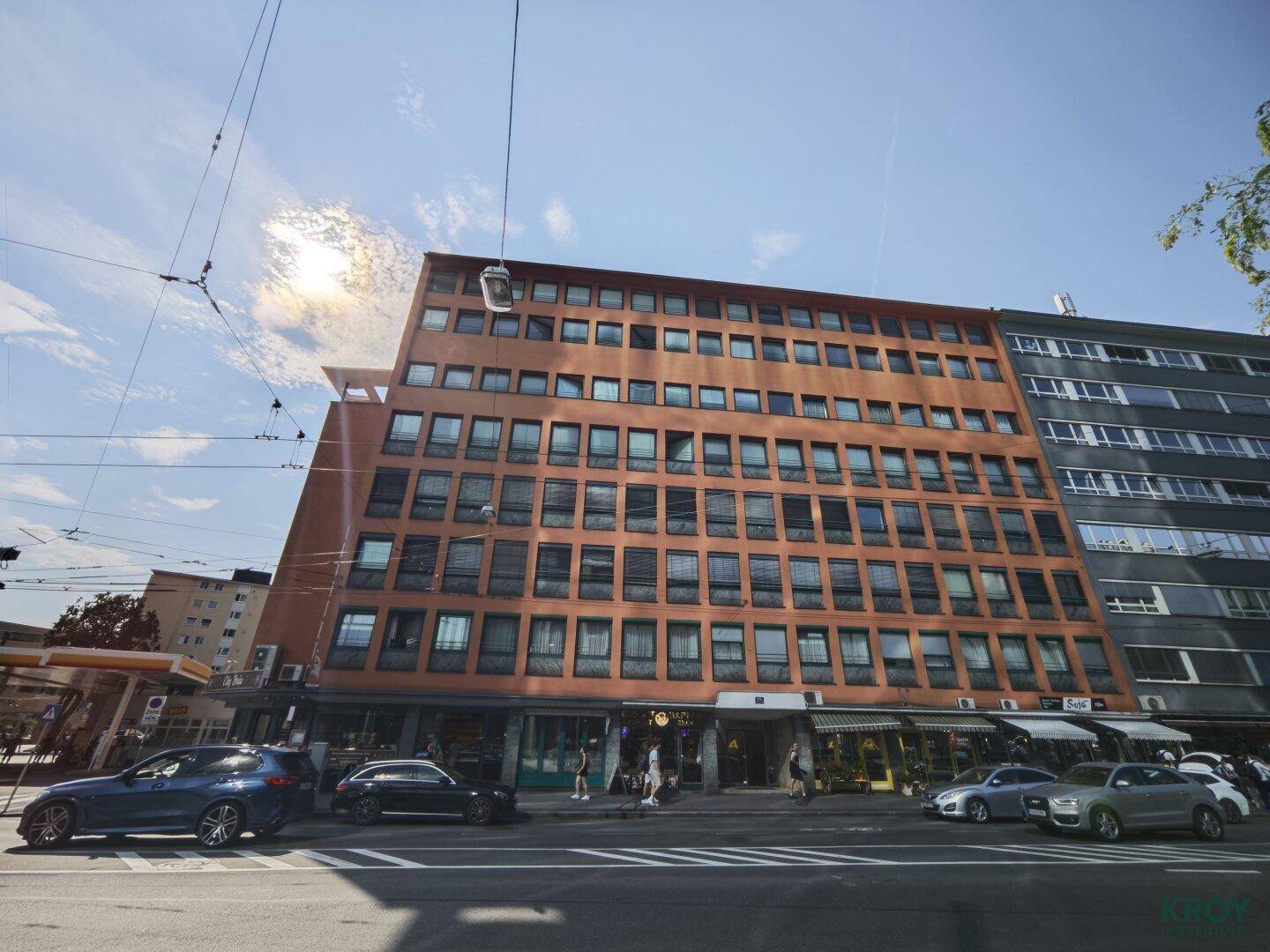 Mehrstöckiges Gebäude mit vielen Fenstern und Geschäften im Erdgeschoss an einer belebten Straße.