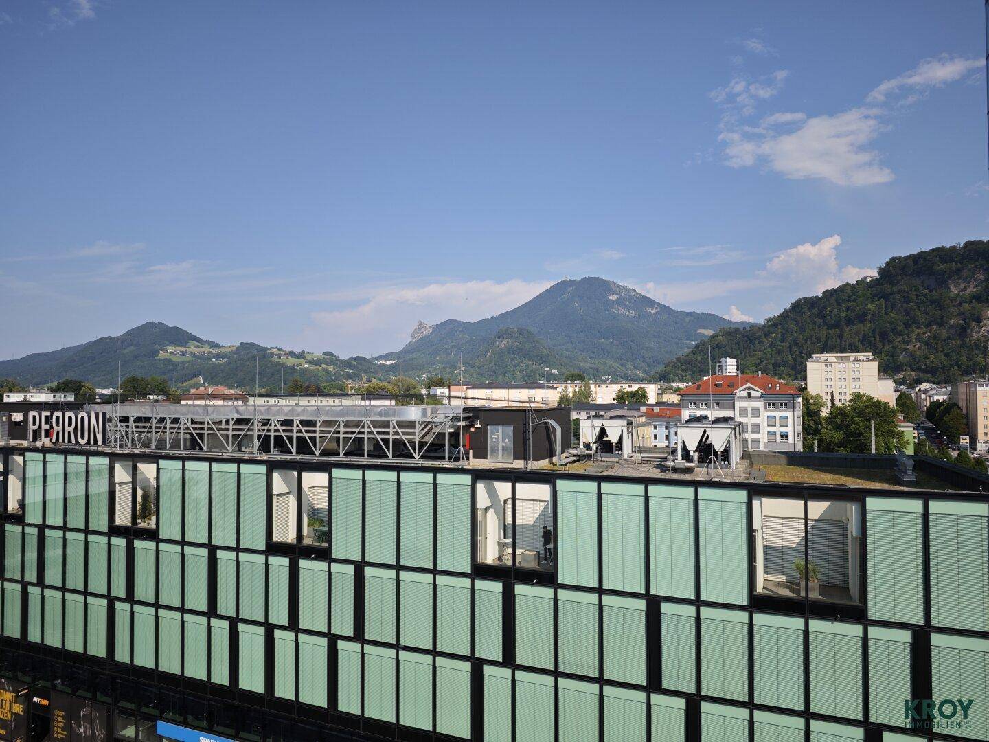 Weitblick über Dächer und moderne Gebäude mit Bergen im Hintergrund unter blauem Himmel.
