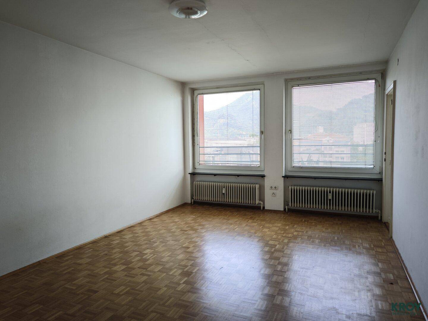 Leeres Wohnzimmer mit Parkettboden, zwei Fenstern und Blick auf die Berge.