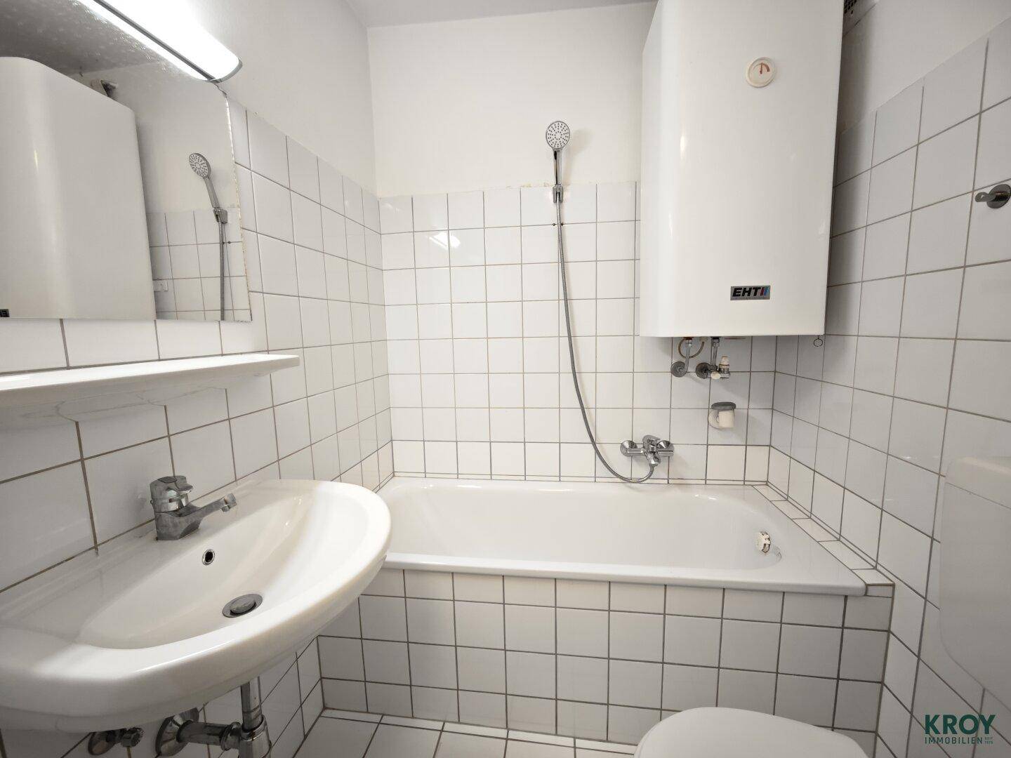 Gefliestes Badezimmer mit Badewanne, Waschbecken und einem Warmwasserboiler an der Wand.