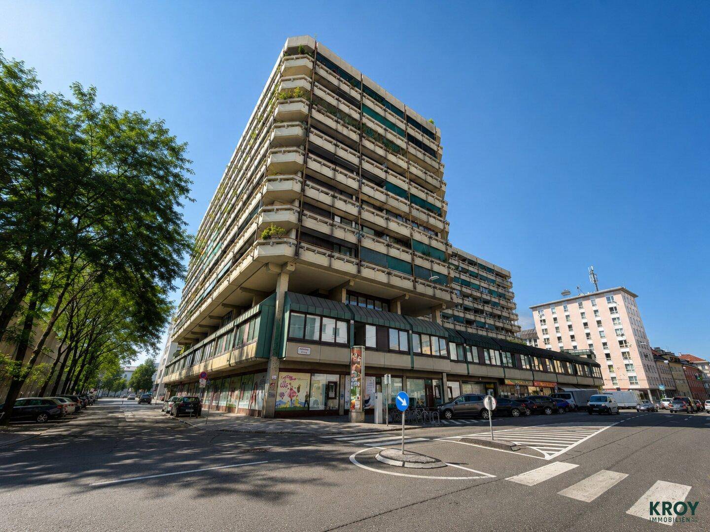 Mehrstöckiges Wohn- und Geschäftshaus mit zahlreichen Balkonen und Geschäften im Erdgeschoss an einer baumgesäumten Straße.