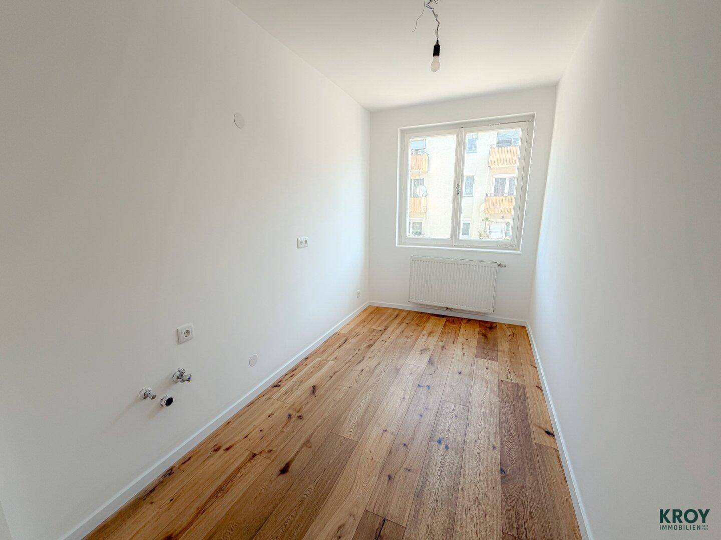 Kleiner Raum mit Holzboden und Fenster, vorbereitet für Installationen wie Waschmaschine oder Küchenzeile.