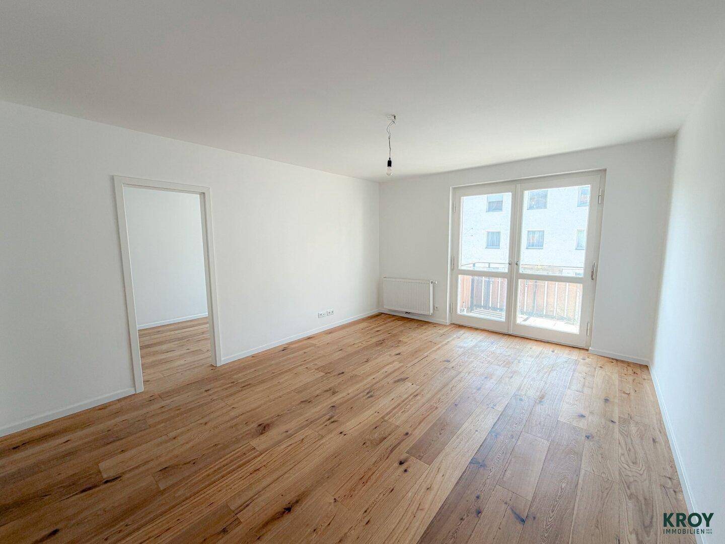 Geräumiges Zimmer mit hellem Holzboden und Zugang zu weiteren Räumen sowie zum Balkon.