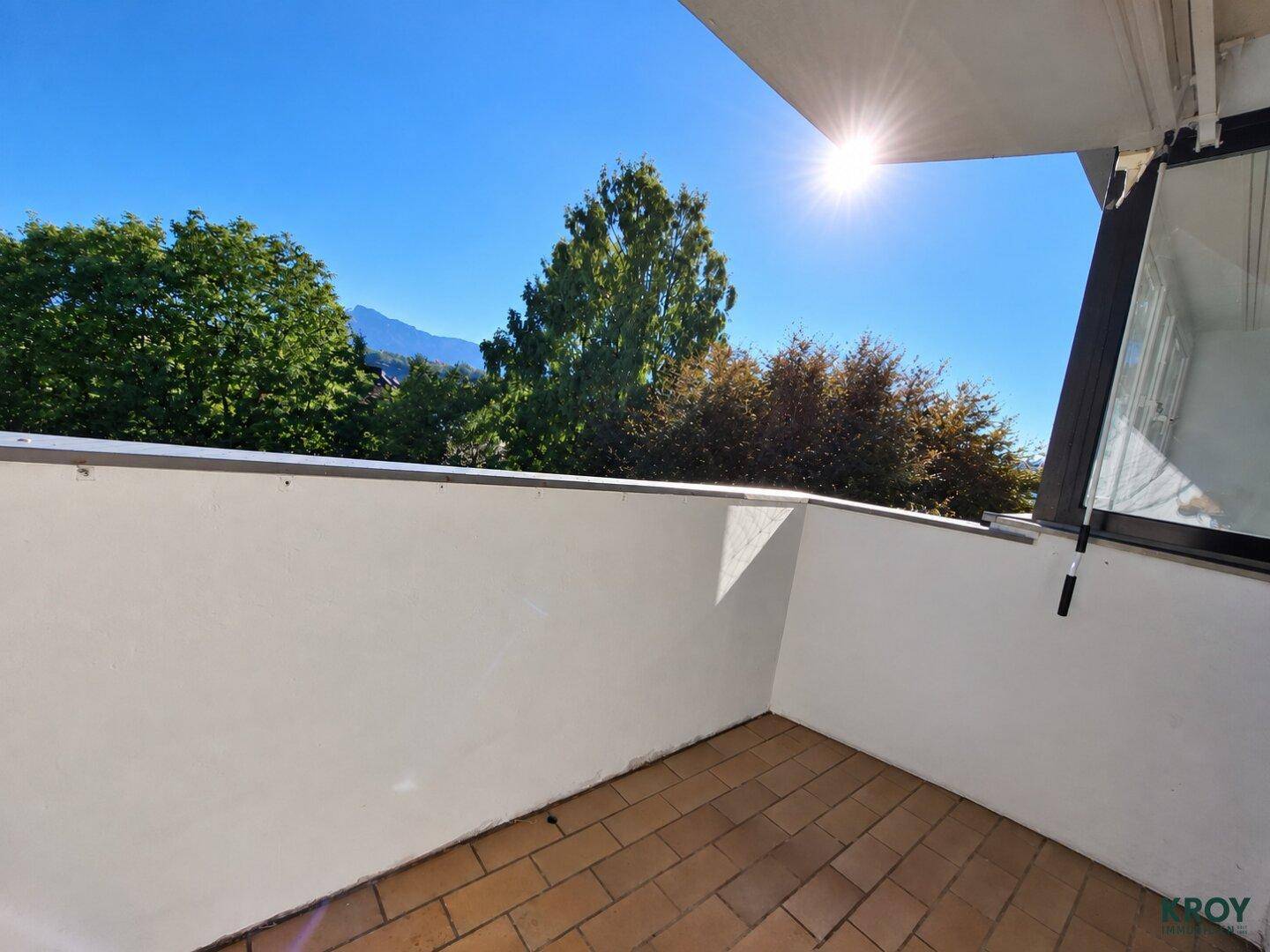 Sonniger Balkon mit Fliesenboden und Blick auf grüne Bäume und die umliegende Berglandschaft.