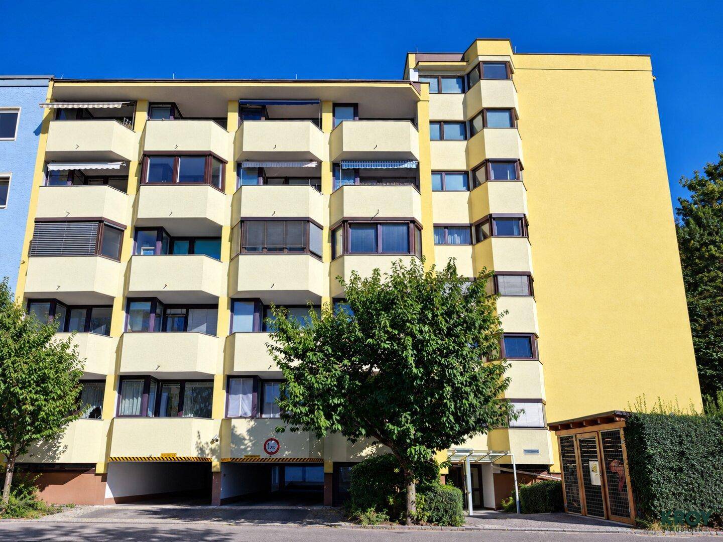 Modernes Mehrfamilienhaus mit gelber Fassade, vielen Balkonen und Tiefgaragenzufahrt.