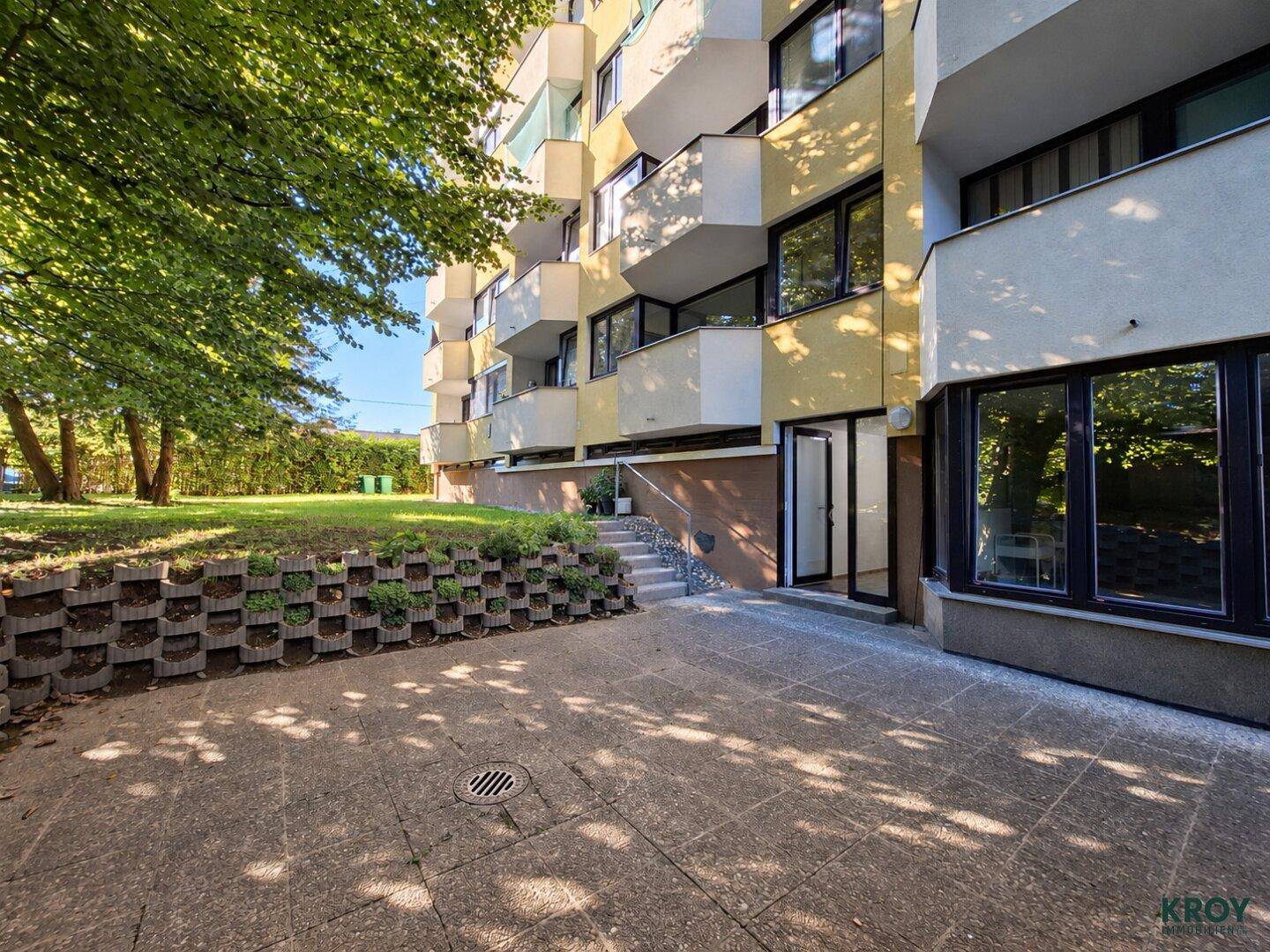 Außenbereich mit gepflasterter Terrasse und Zugang zu einem grünen Garten neben dem Gebäude.
