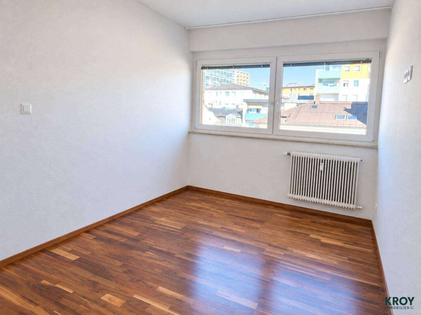 Helles Schlafzimmer mit Parkettboden und Blick auf die umliegenden Gebäude und den Himmel.