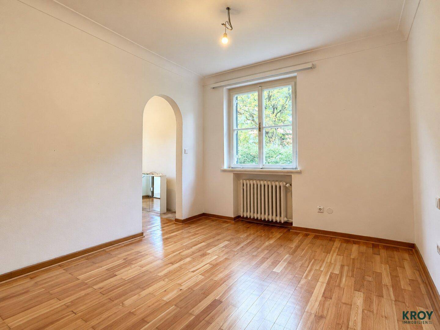 Geräumiges Zimmer mit Parkettboden, weiß gestrichenen Wänden und einem Bogen zu einem Nebenraum.