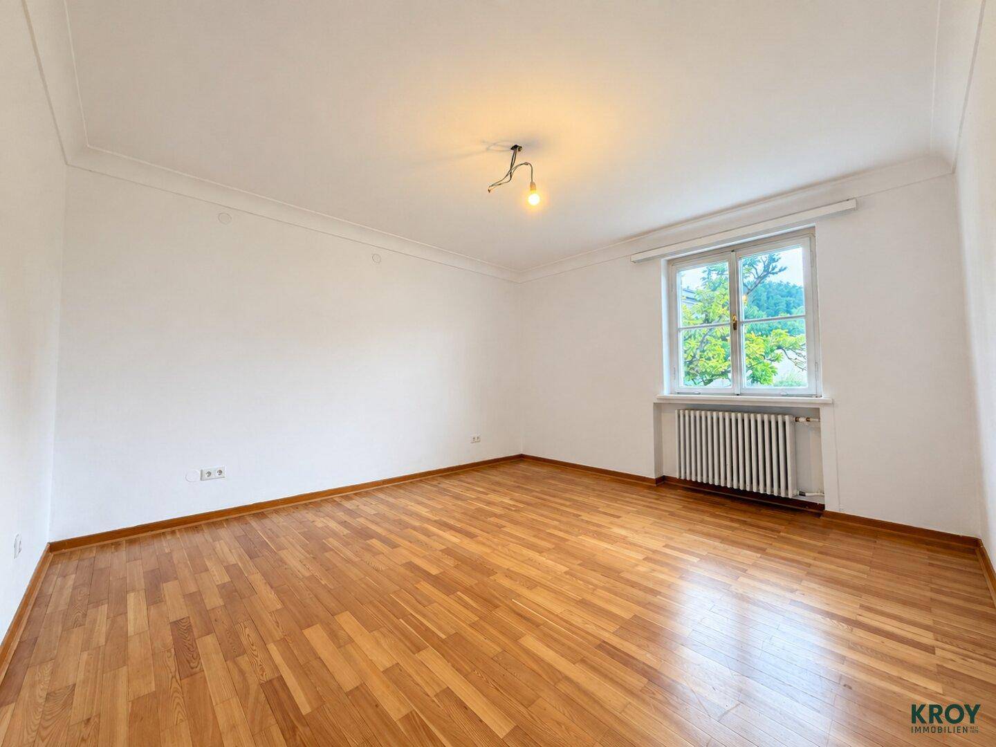 Ruhiges Schlafzimmer mit Parkettboden, weißen Wänden und einem Fenster mit Blick nach draußen.