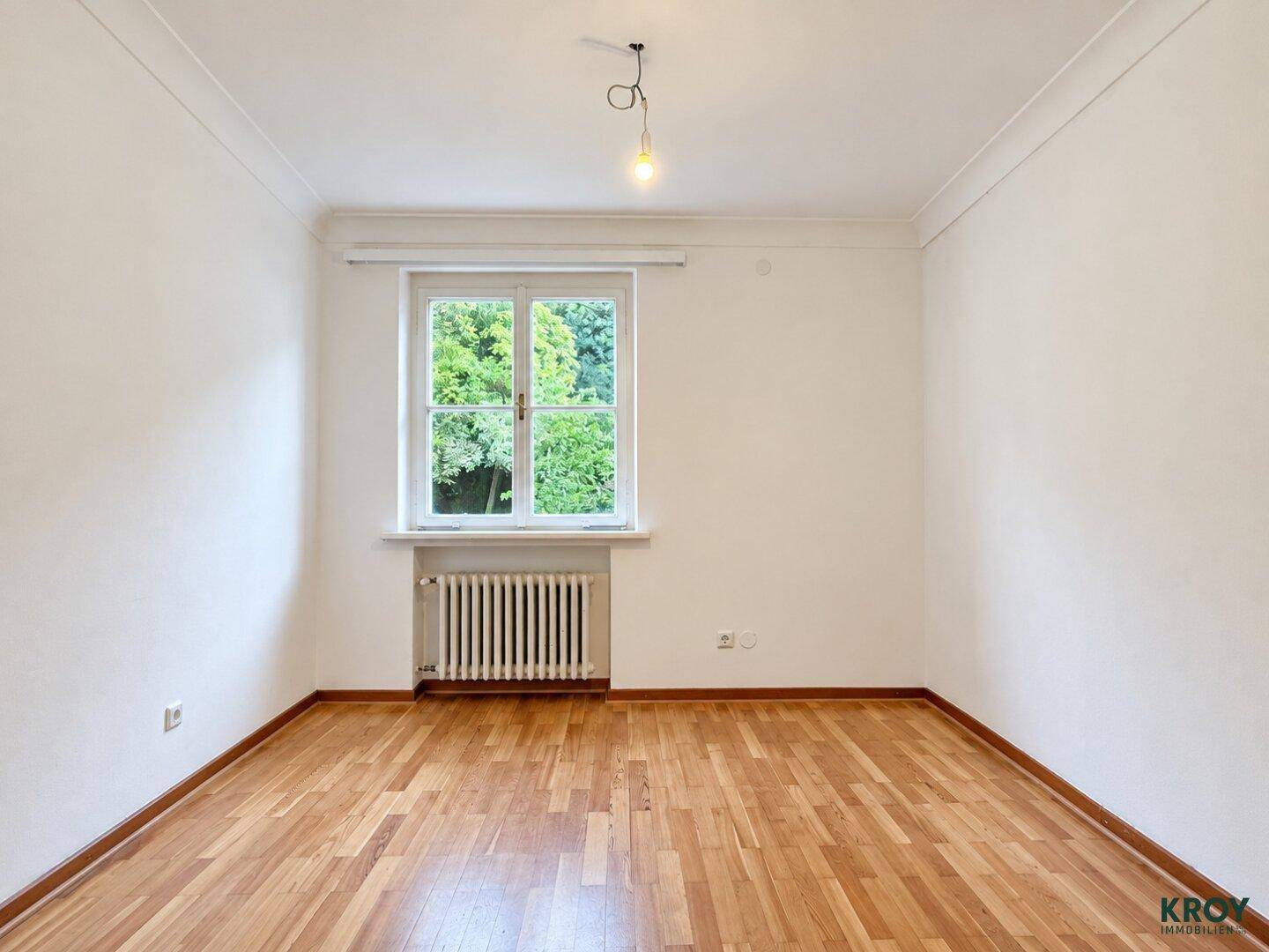 Gemütliches Zimmer mit Parkettboden, weiß gestrichenen Wänden und einem großen Fenster.