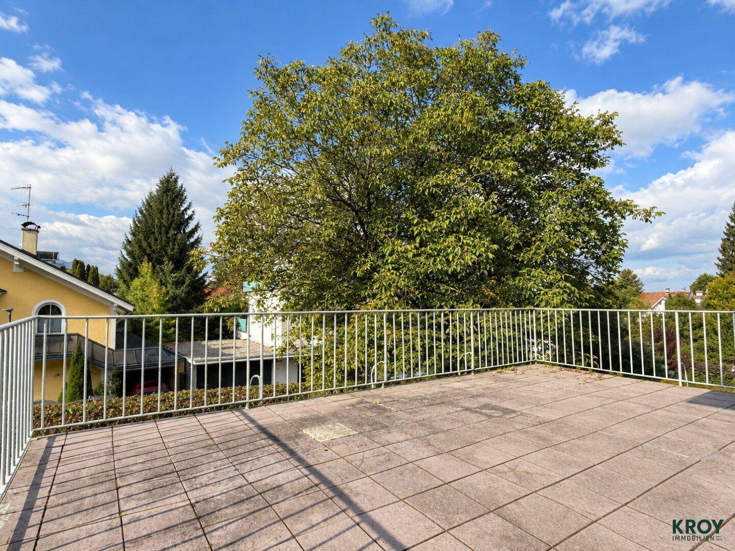 Weitläufige Terrasse mit Fliesenboden und weitem Blick auf grüne Bäume und blauen Himmel.