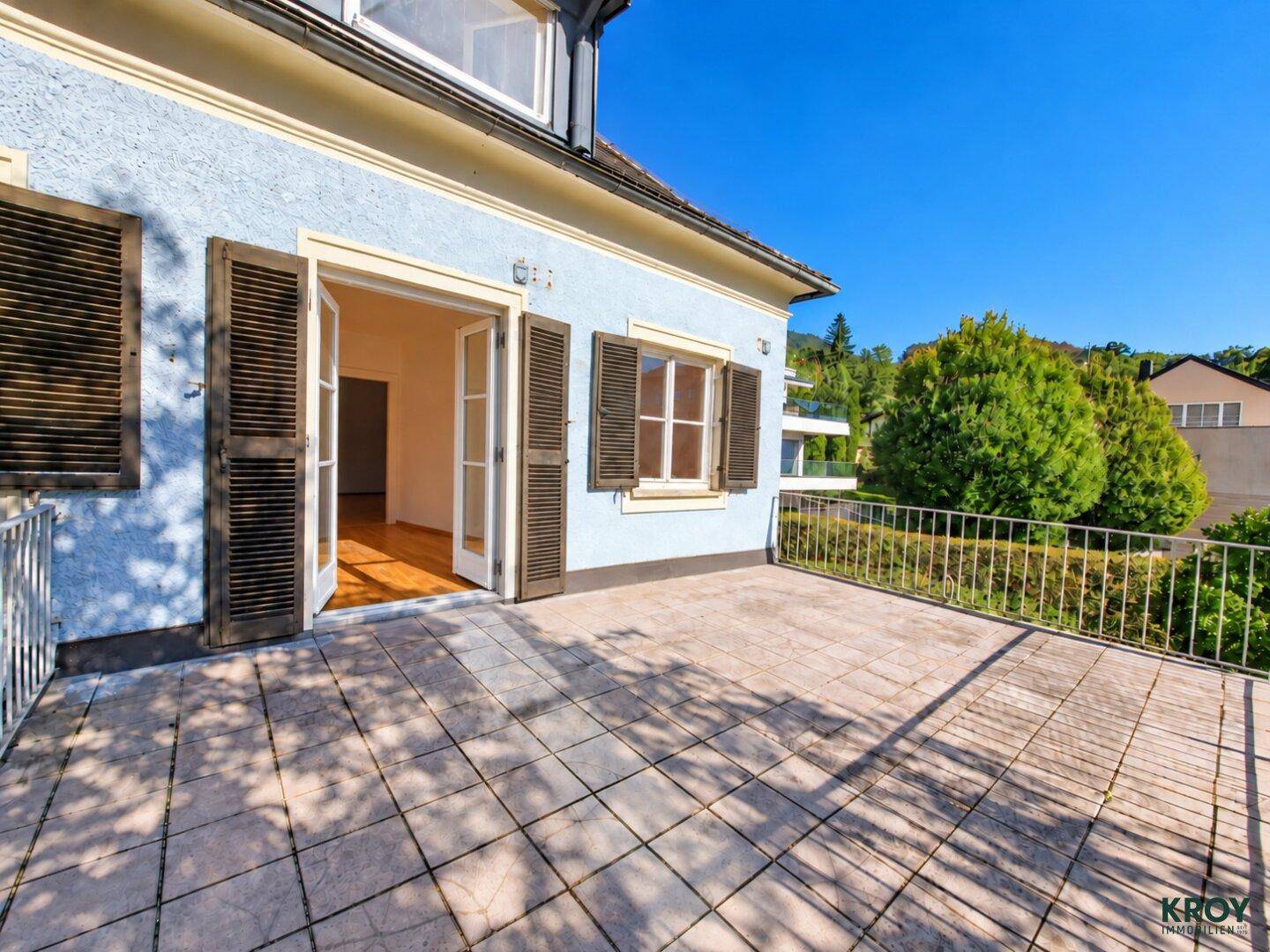 Geräumige Terrasse mit Fliesenboden und Zugang zu einem hellblauen Haus mit Fensterläden.