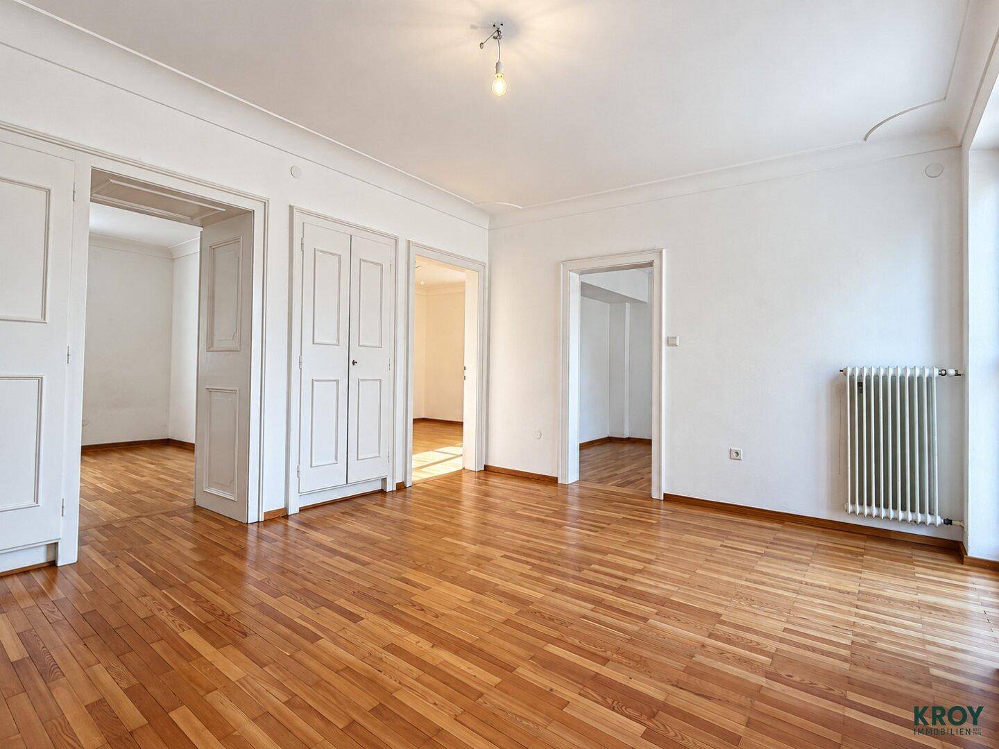 Helles Zimmer mit Parkettboden, weißen Wänden und mehreren Türen zu angrenzenden Räumen.