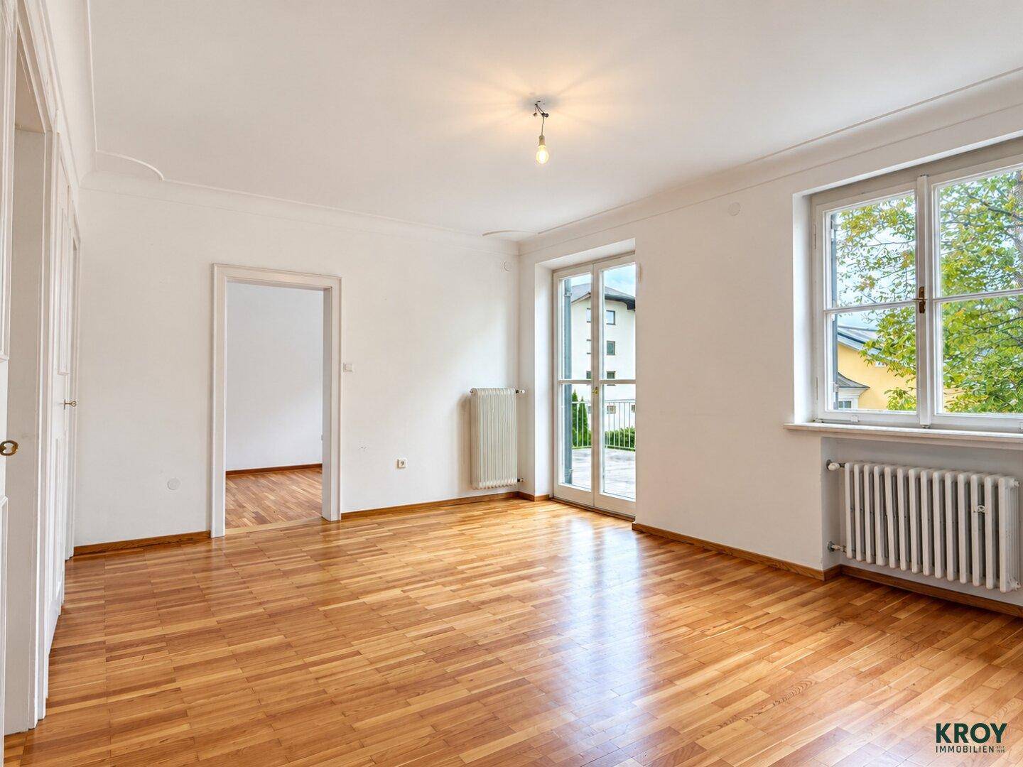 Heller Raum mit Parkettboden, Fenster und Zugang zu einem weiteren Zimmer mit Balkon.