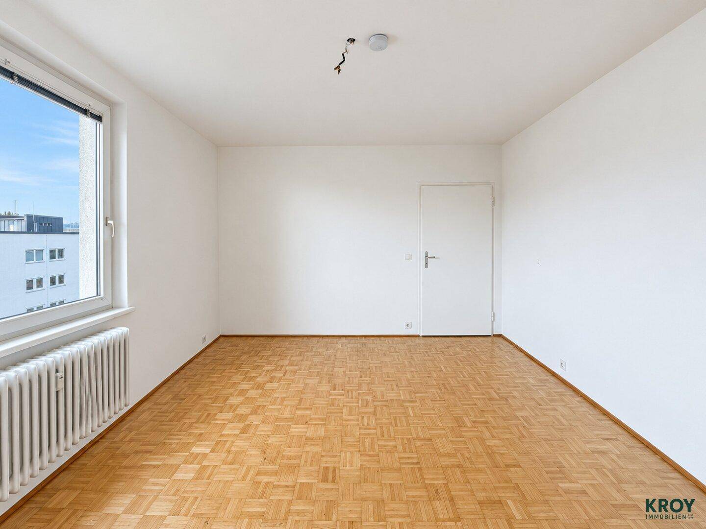 Geräumiges, leeres Zimmer mit Parkettboden und weiß gestrichenen Wänden.