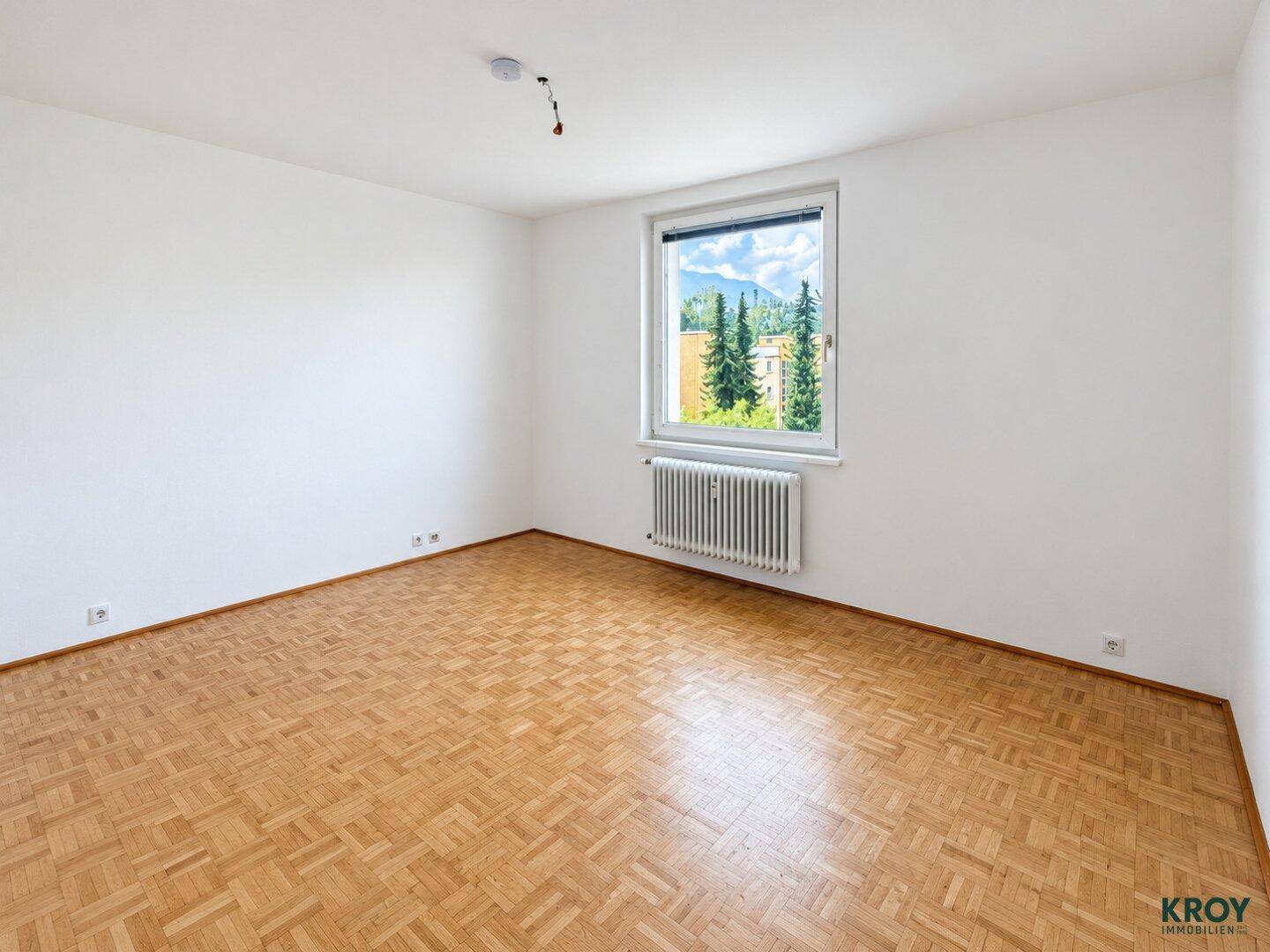 Helles, leeres Zimmer mit Parkettboden und Fenster mit Blick ins Grüne.