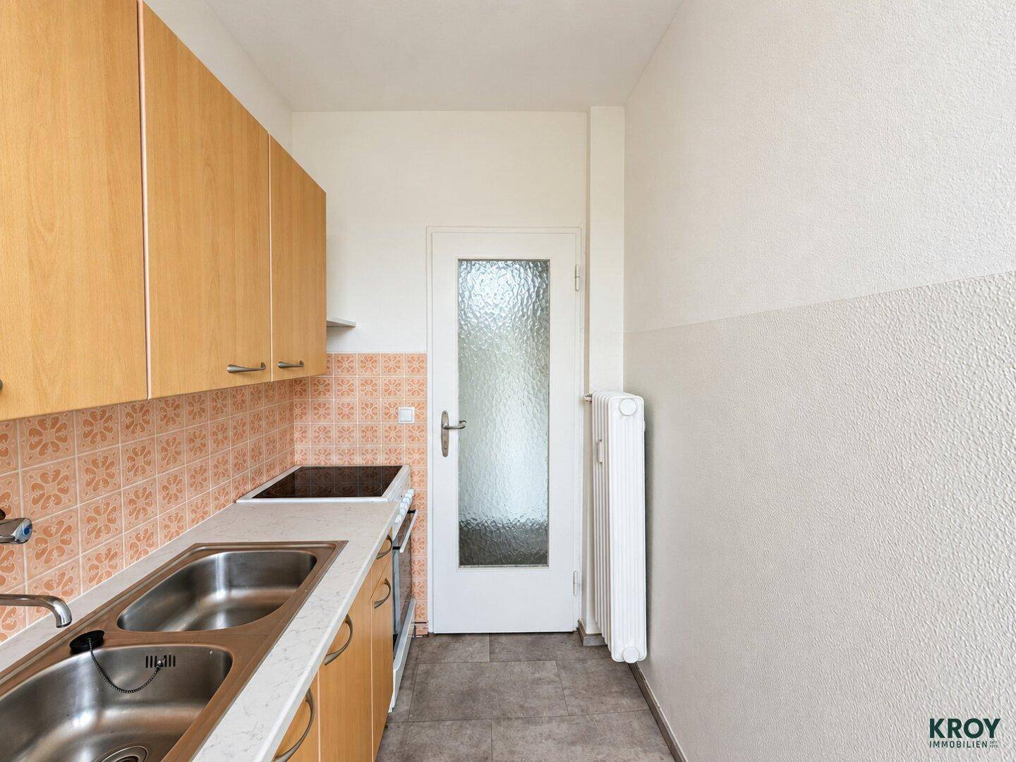Schmale Küche mit Holzschränken, Fliesenboden und Heizkörper.