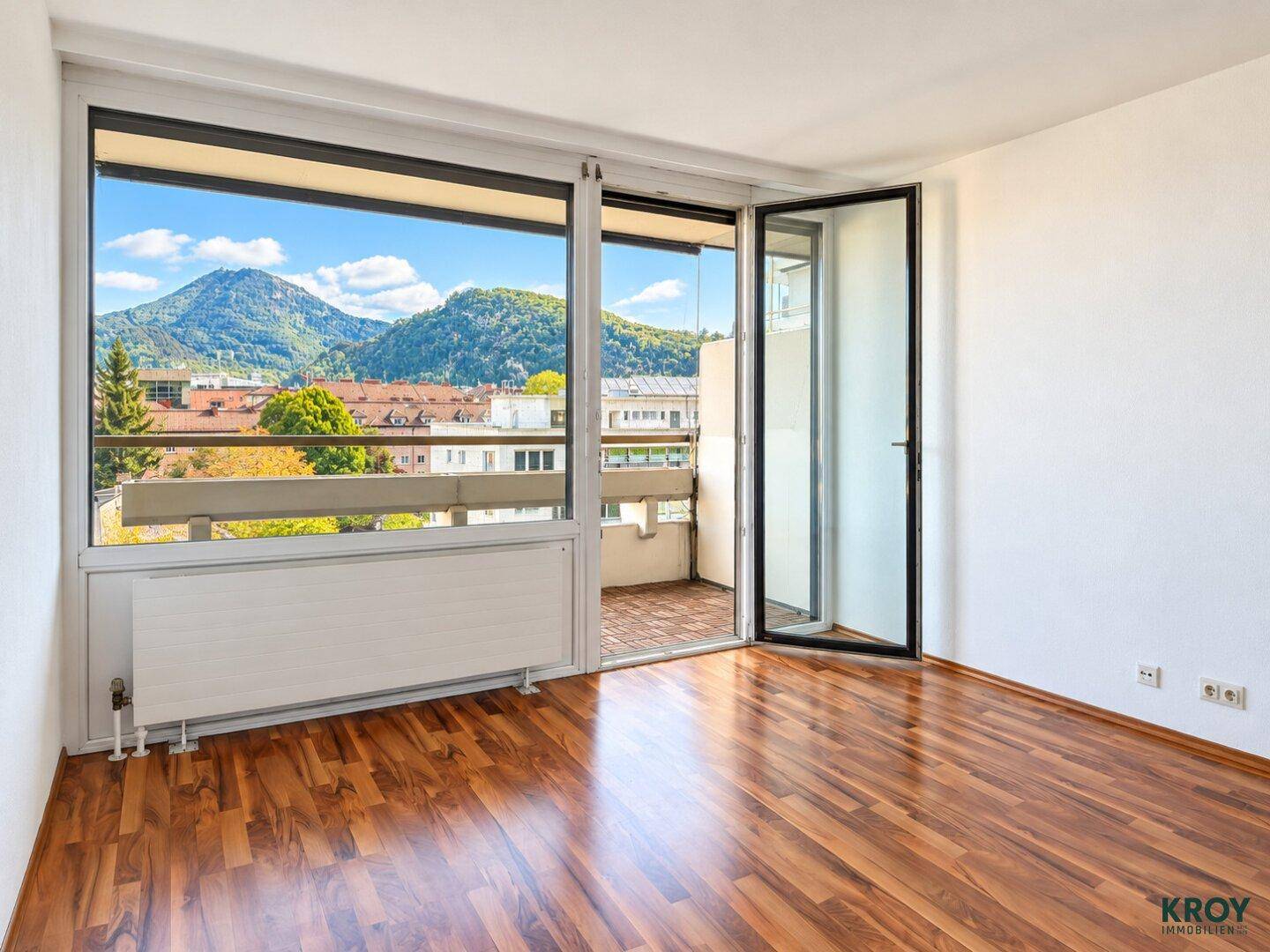 Lichtdurchflutetes Zimmer mit Holzboden und Balkonzugang, mit schöner Aussicht auf die umliegende Berglandschaft.