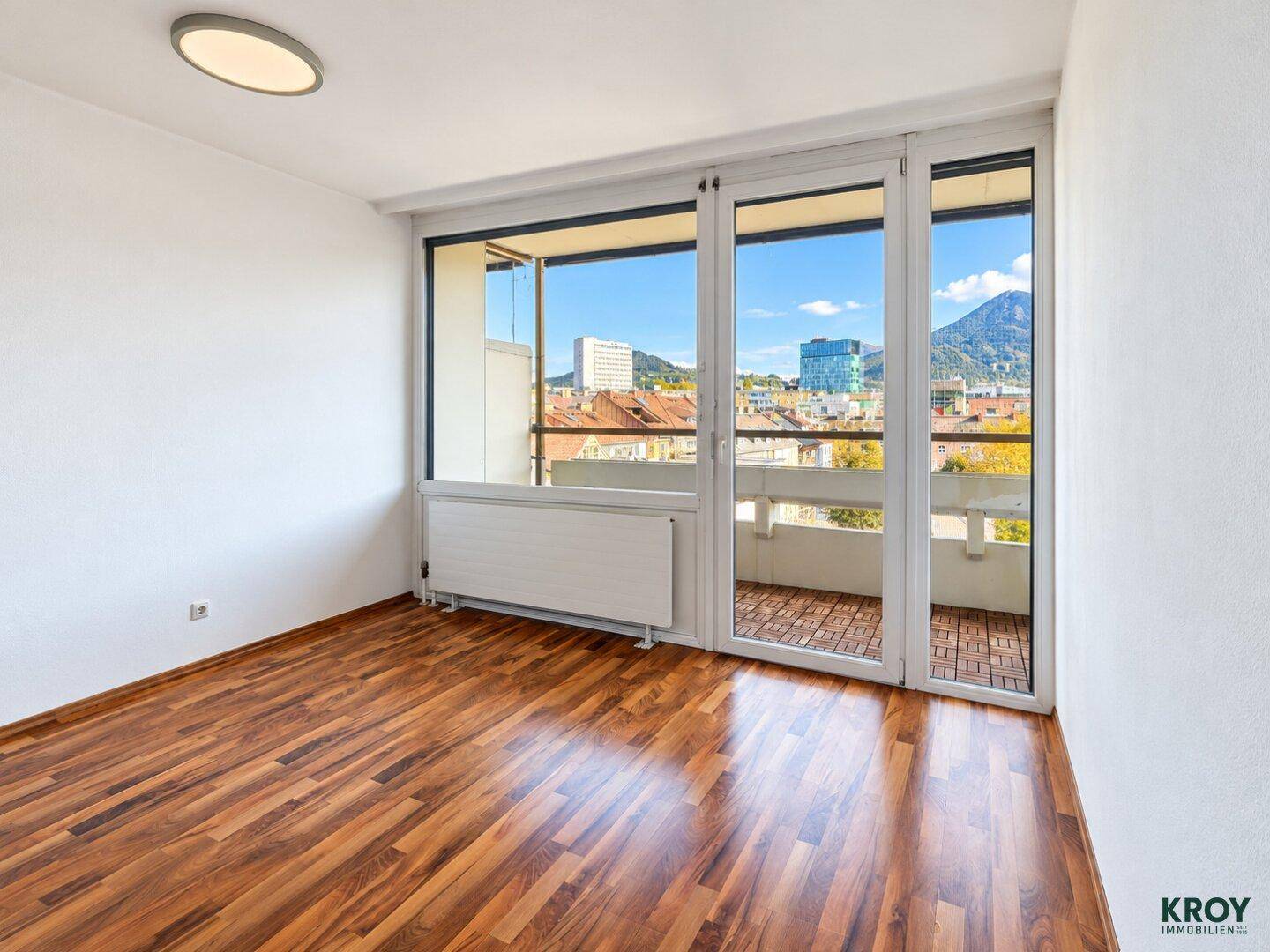Geräumiges Zimmer mit Holzboden und großem Fenster, das einen Blick auf die Stadt und Berge bietet.