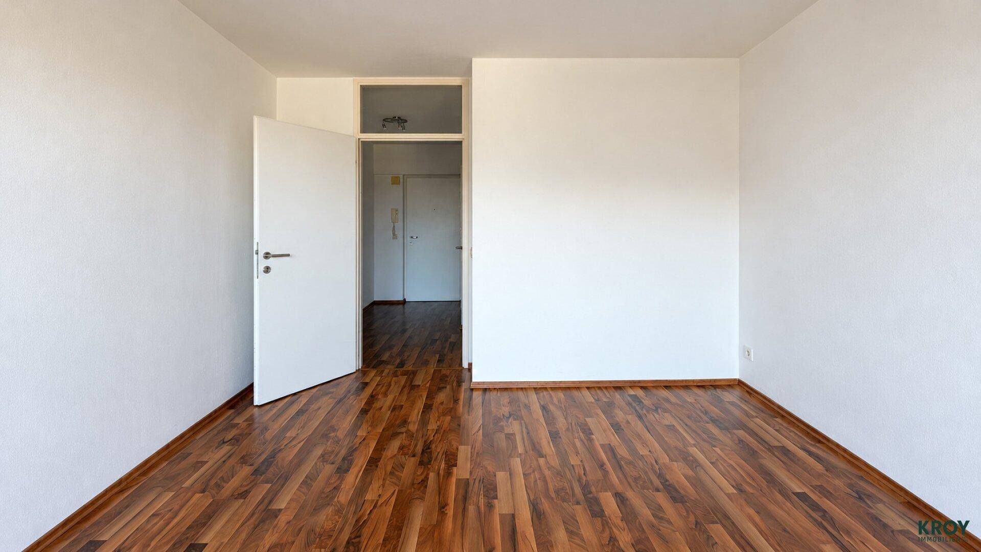 Helles Zimmer mit Holzboden und weiß gestrichenen Wänden, bietet Zugang zu einem weiteren Raum.