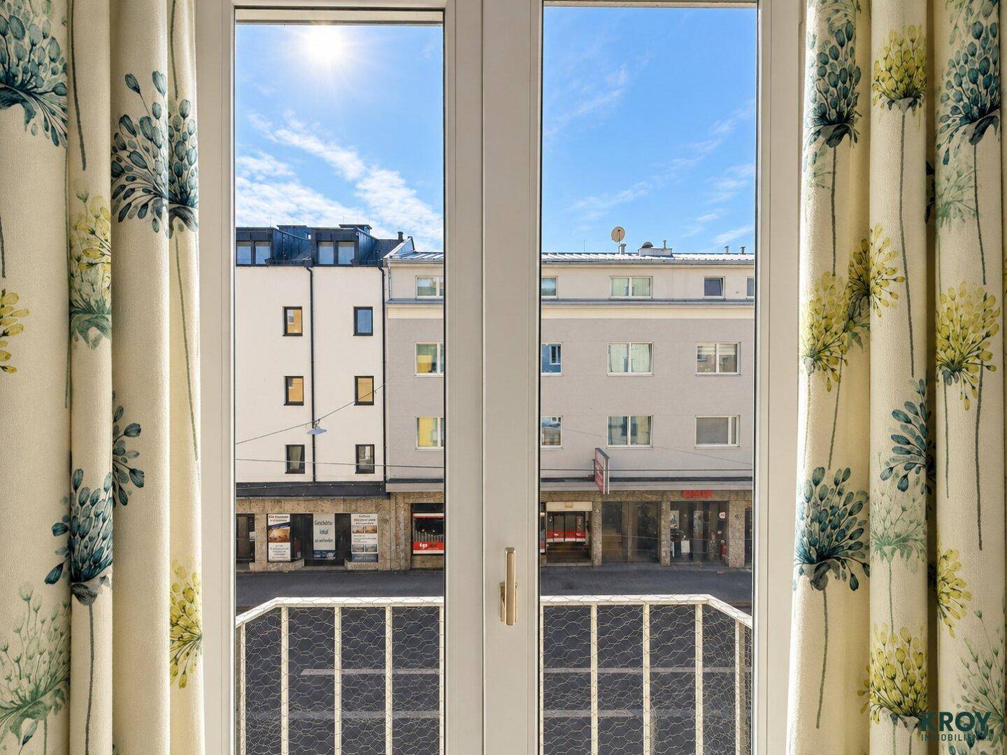 Blick aus dem Fenster auf die gegenüberliegenden Gebäude und die sonnige Straße durch gemusterte Vorhänge.