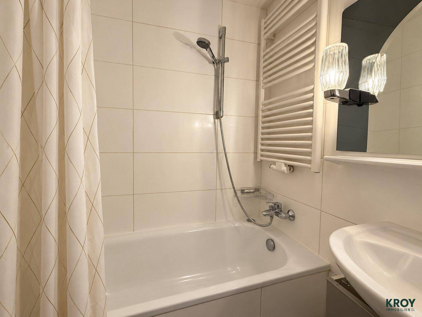 Helles Badezimmer mit Badewanne, Duschvorhang, Handtuchheizkörper und weiß gefliesten Wänden.