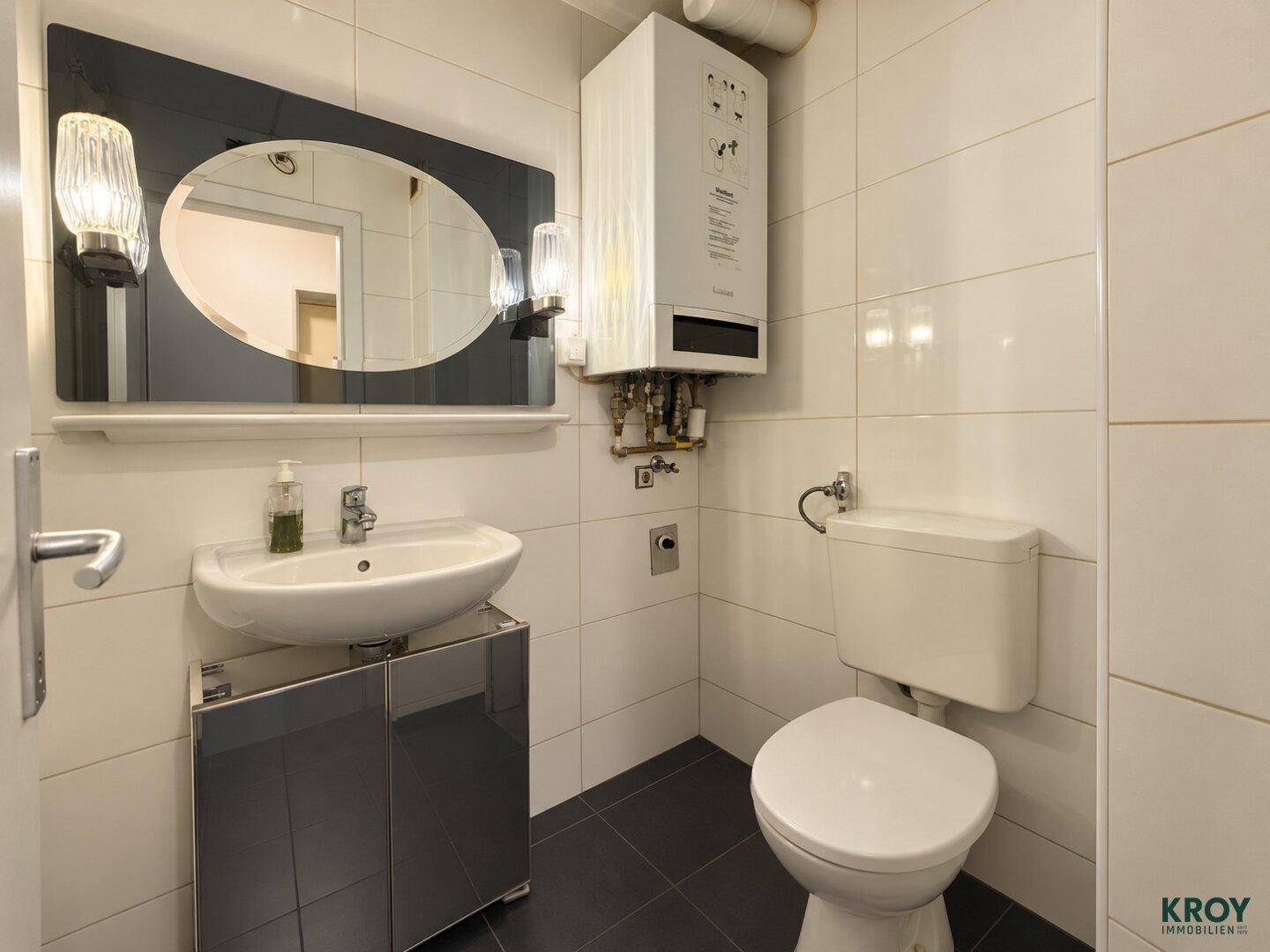 Kleines WC mit Waschbecken, Toilette und Wandspiegel, ausgestattet mit einem Warmwasserboiler.