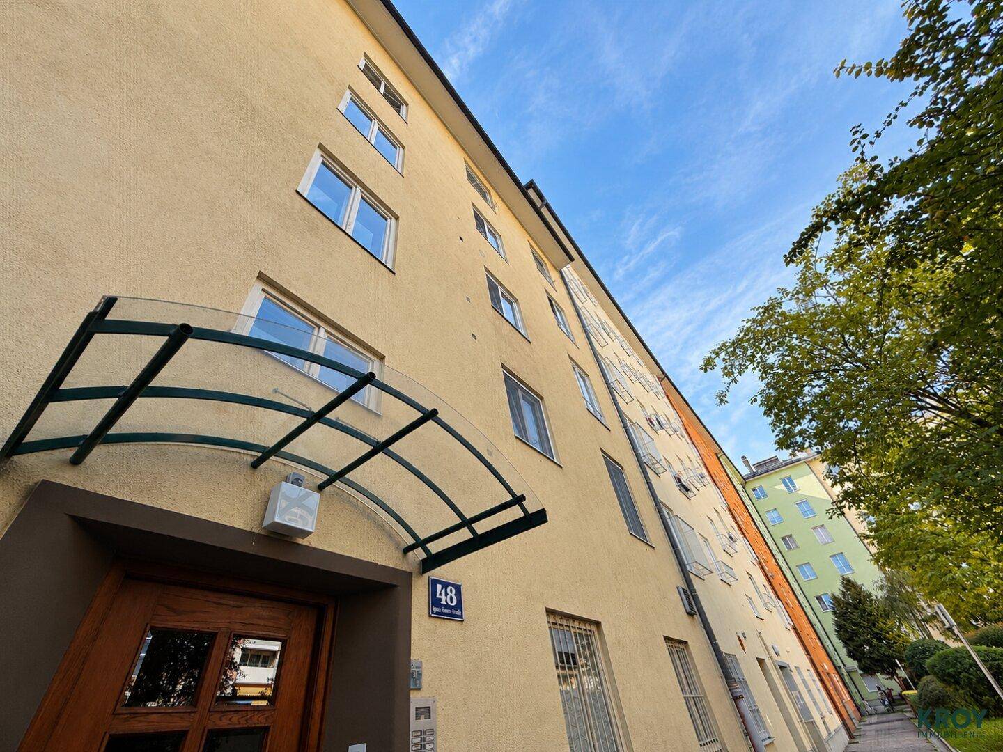 Eingangsbereich eines Mehrfamilienhauses mit gelber Fassade und einem schützenden Glasvordach über der Tür.