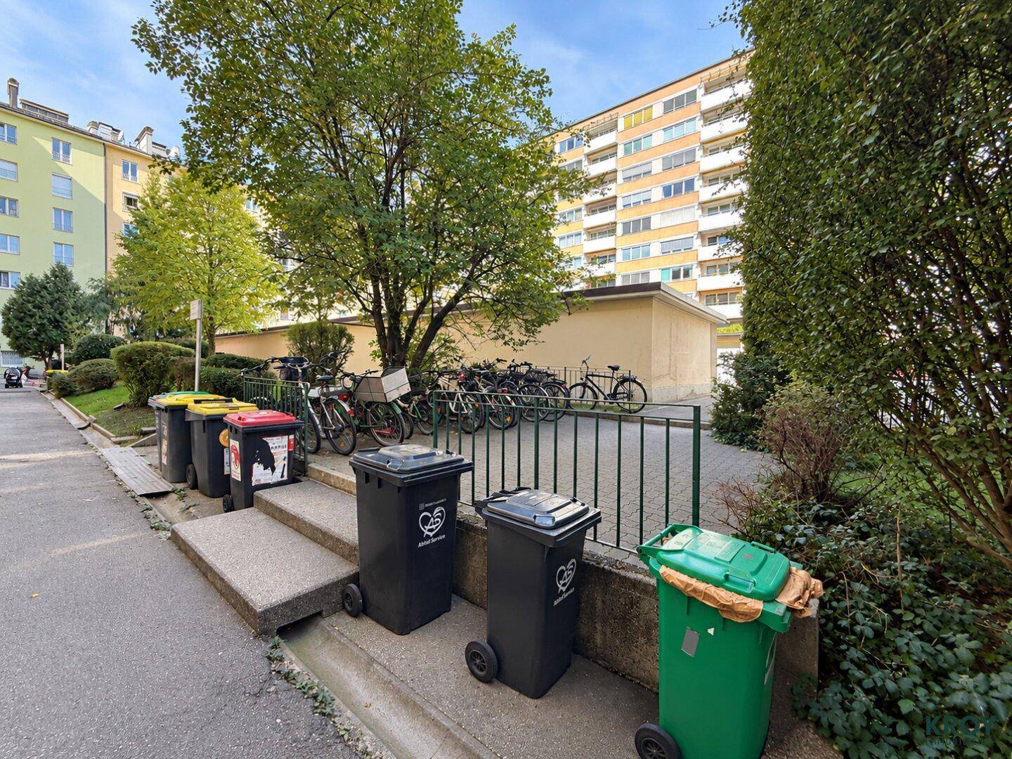 Außenbereich mit Mülltonnen, Fahrradständern und Bäumen, umgeben von weiteren Wohngebäuden und Grünflächen.