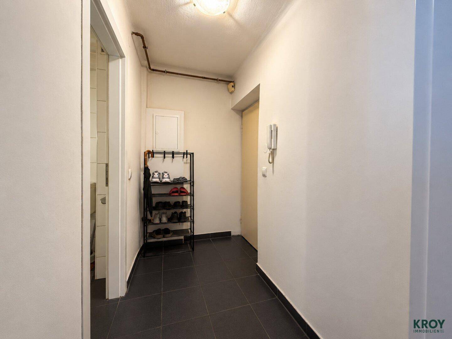 Schmaler Flur mit dunklen Fliesen, Schuhregal und Gegensprechanlage im Eingangsbereich der Wohnung.
