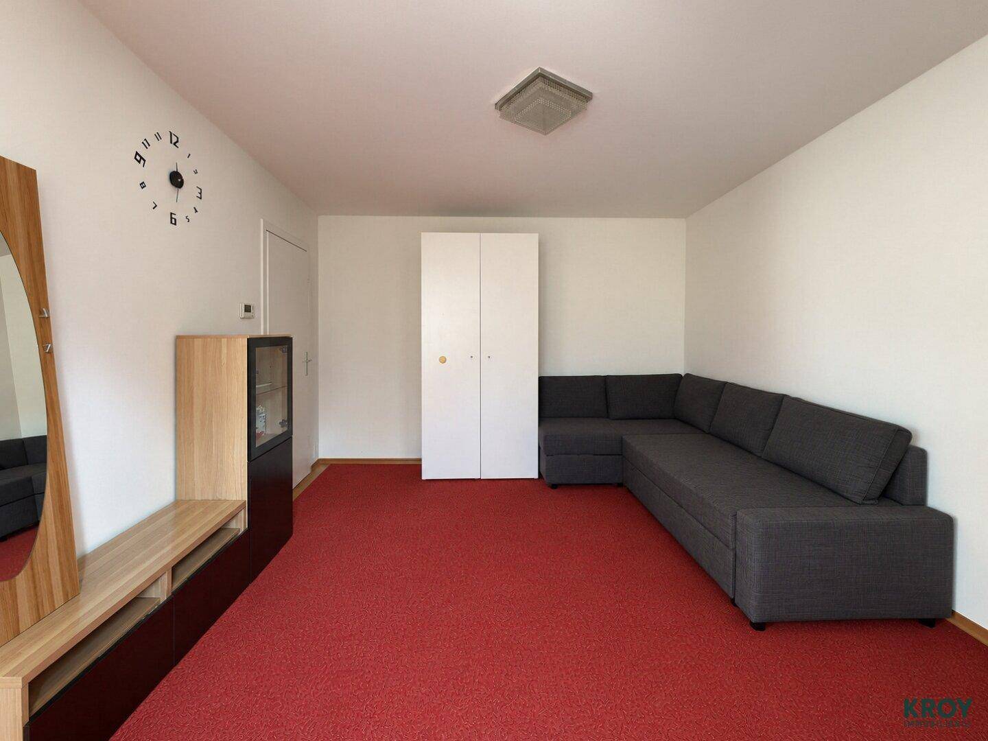 Gemütlicher Wohnraum mit rotem Teppichboden, dunklem Ecksofa und schlichten Holzmöbeln.