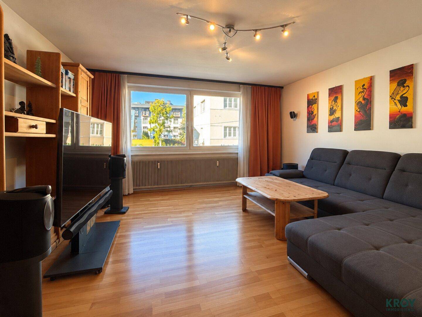 Gemütliches Wohnzimmer mit Holzboden, großem Fenster, grüner Aussicht und bequemer Eckcouch.