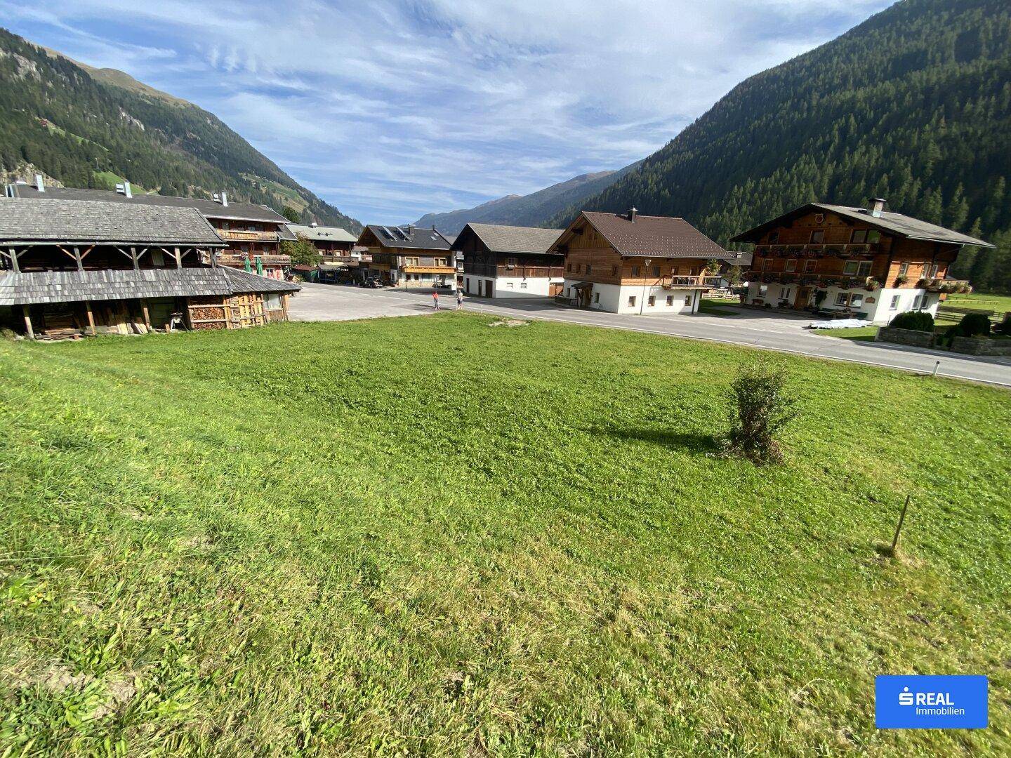 Panoramablick über eine weite Wiese mit traditionellen Holzhäusern und majestätischen Bergen im Tal.
