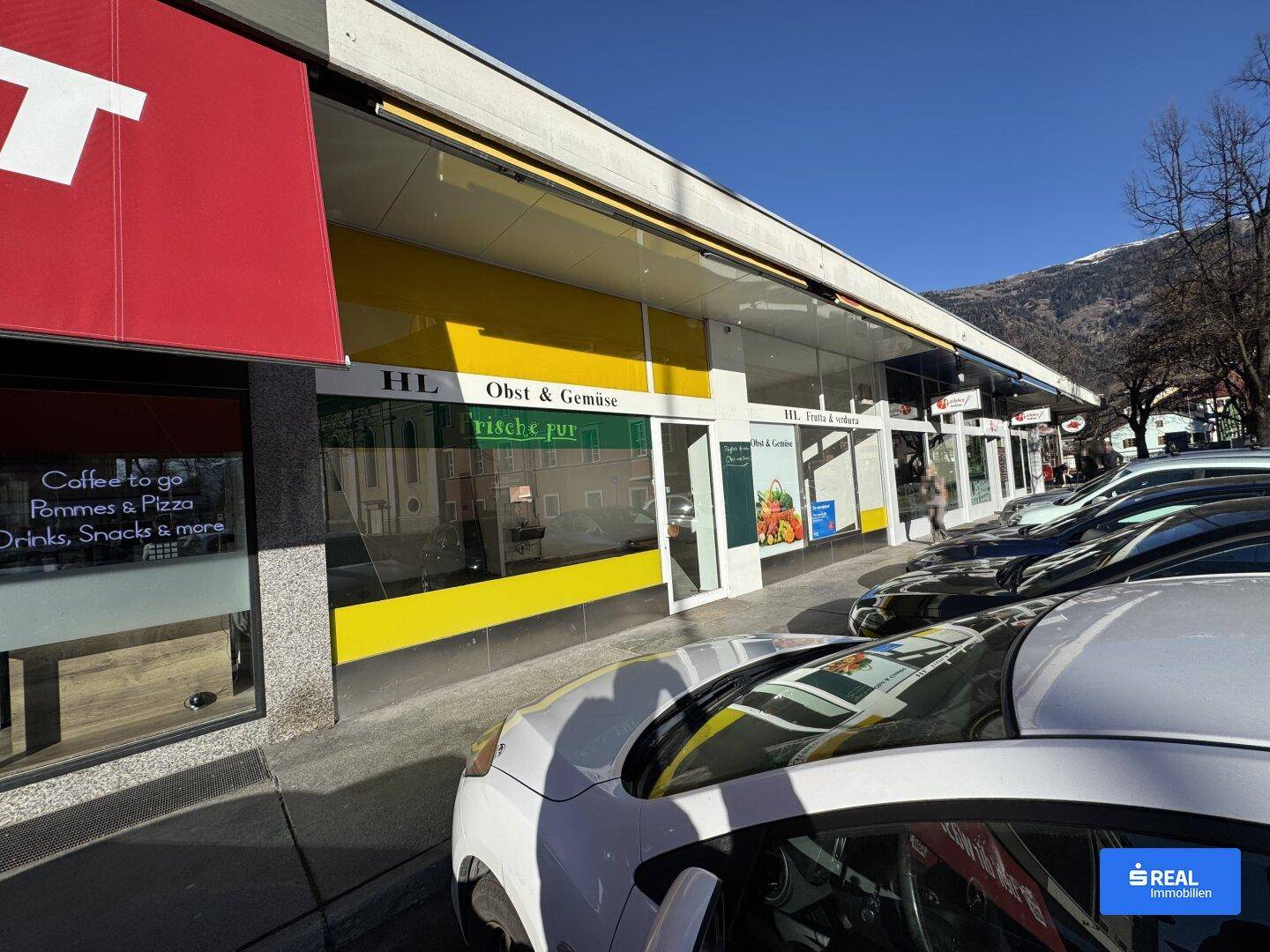 Ansicht der Ladenfronten eines Geschäftsgebäudes mit geparkten Autos und Bergen im Hintergrund an einem sonnigen Tag.