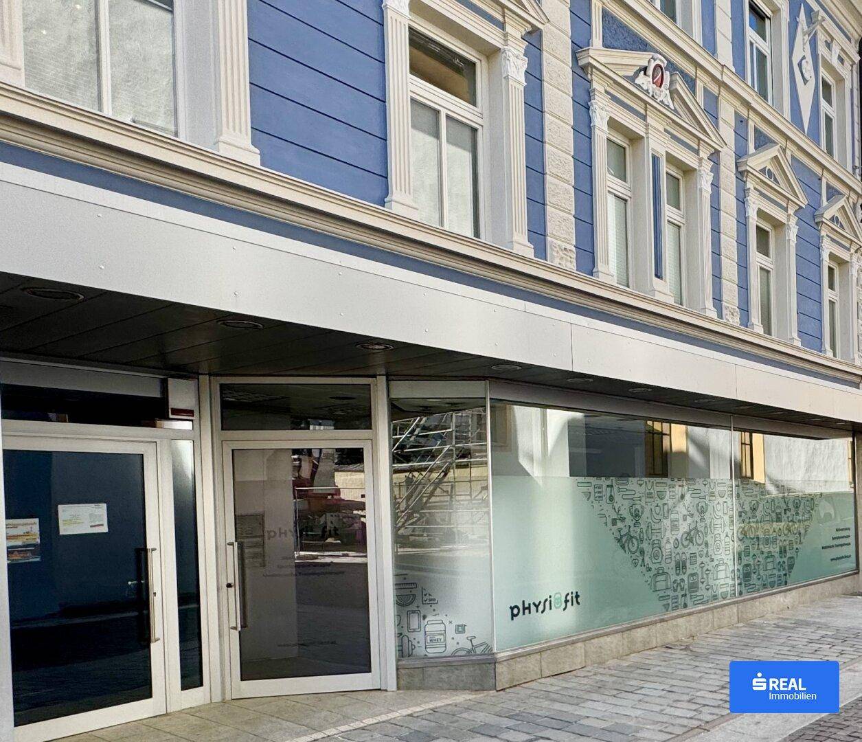 Die Fassade des Gebäudes zeigt eine blaue Verkleidung und große Schaufenster im Erdgeschoss.