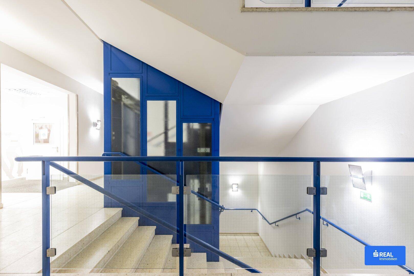 Helles Treppenhaus mit modernem Aufzug, blauen Akzenten und Glasgeländer, das viel natürliches Licht hereinlässt.