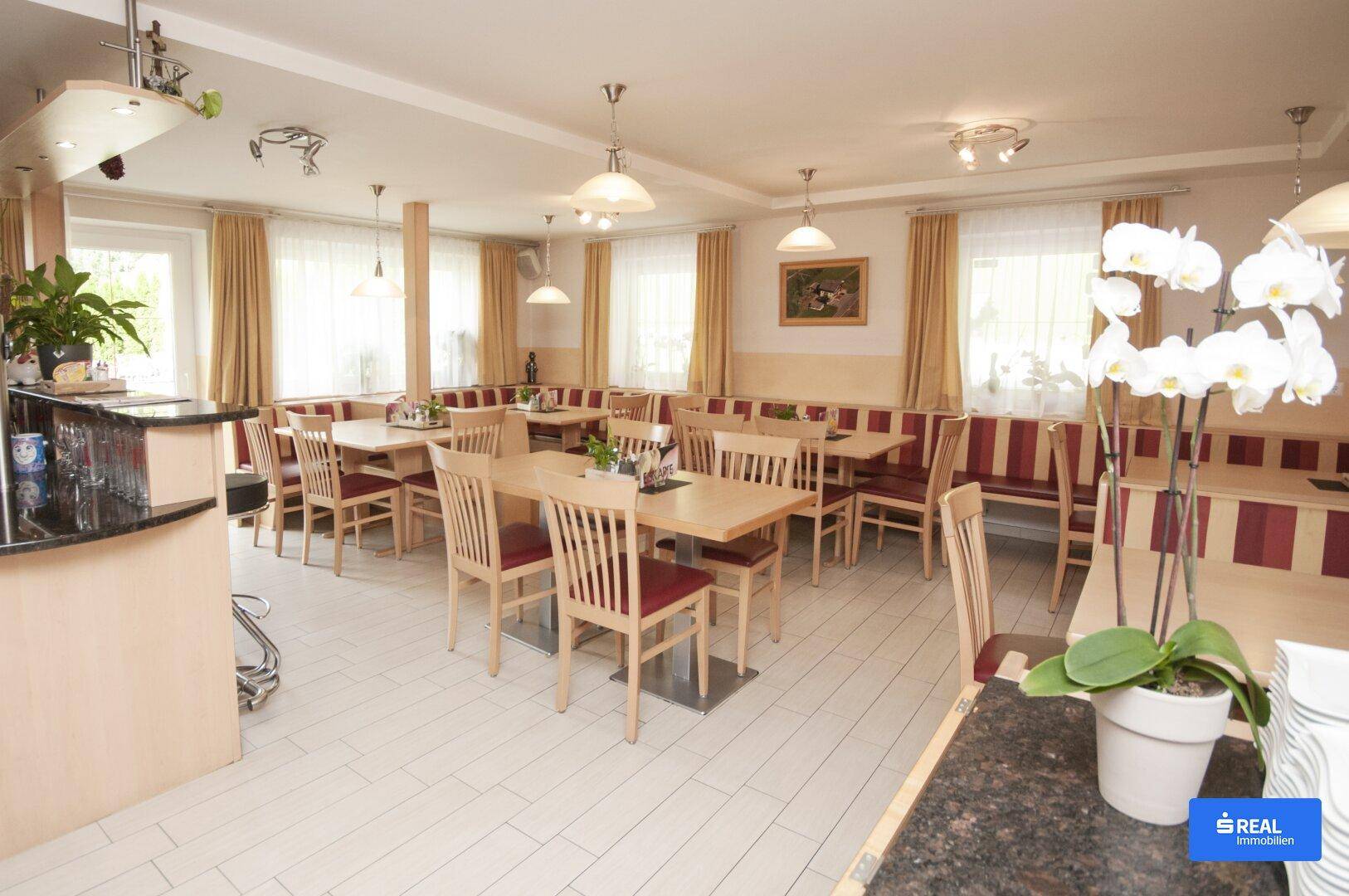 Geräumiger Gastraum mit mehreren Tischen, Stühlen und Sitzbänken, ideal für ein Restaurant oder Café. Helle Fliesen und große Fenster sorgen für eine freundliche Atmosphäre.
