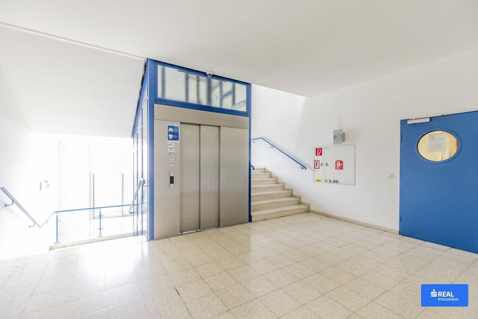 Heller Flur mit modernem Aufzug, Treppenhaus und blauer Tür, der Zugang zu den Räumlichkeiten bietet.