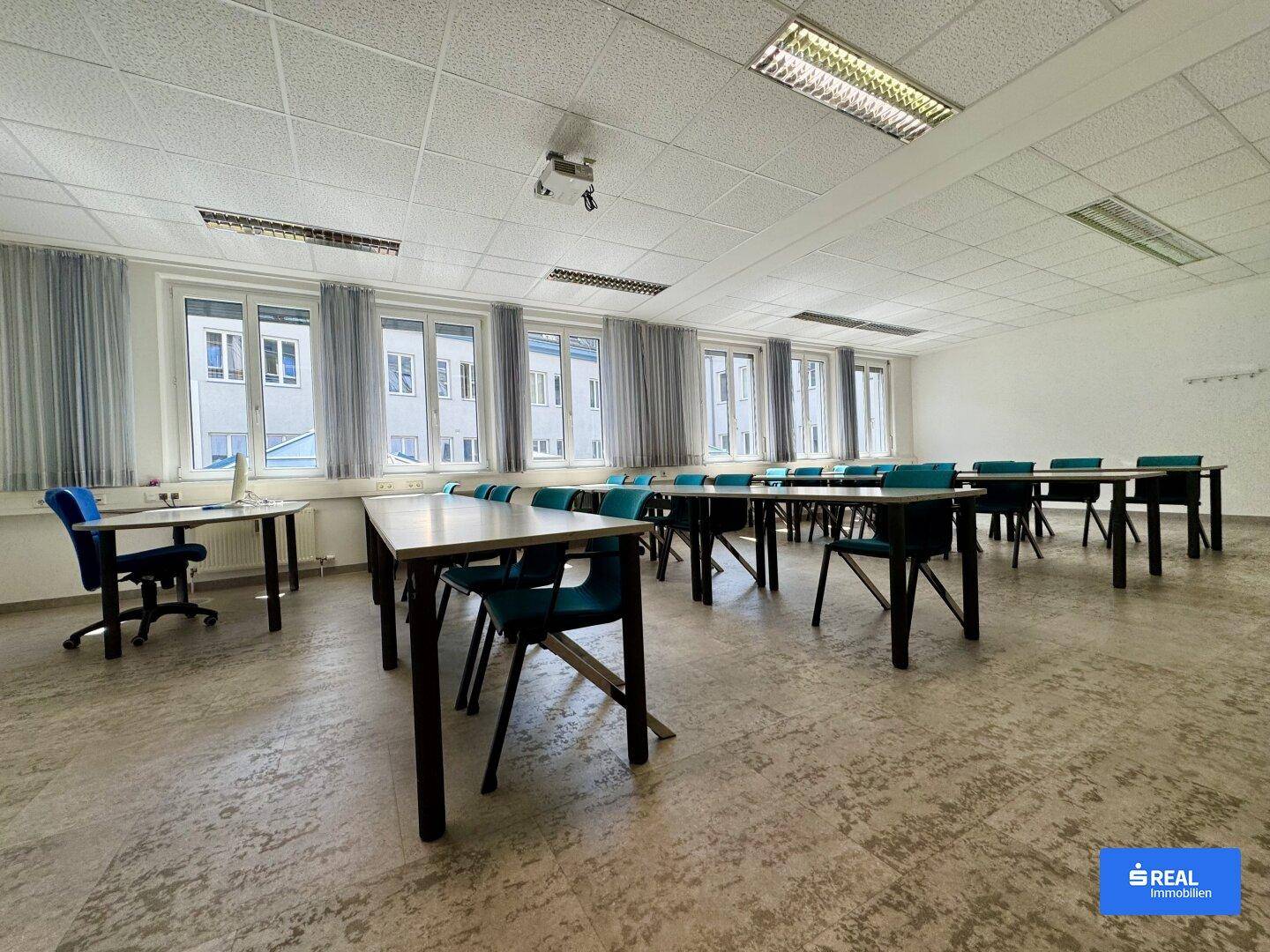 Großer Konferenzraum mit zahlreichen Tischen und Stühlen, hellen Fenstern und Blick auf die umliegenden Gebäude.