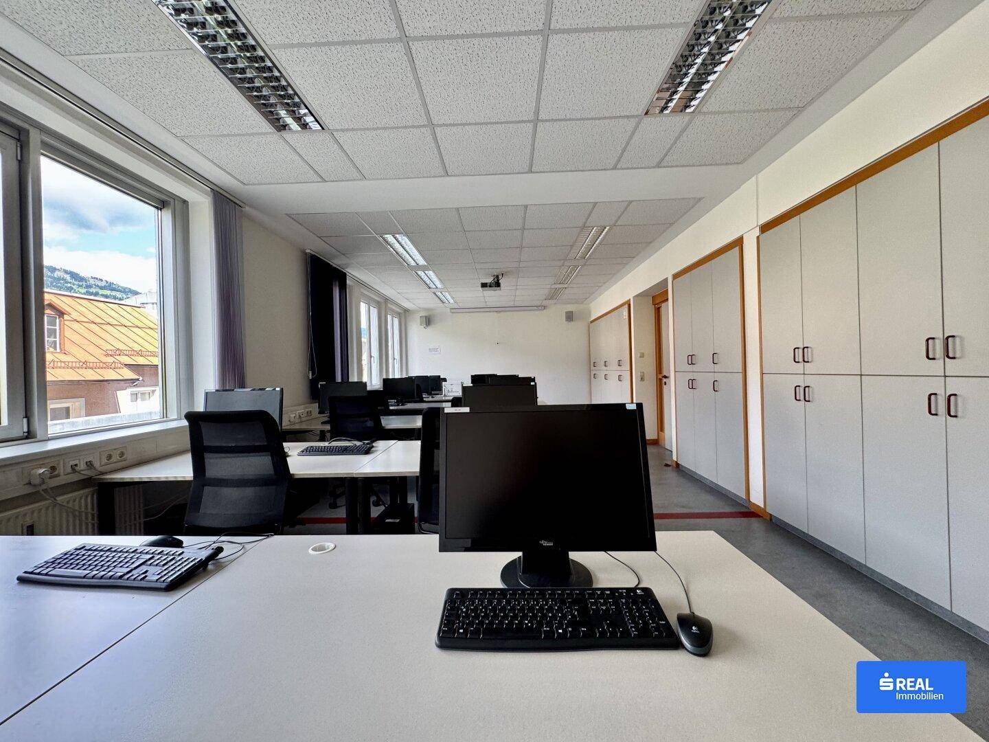 Büroraum mit Computern, Schreibtischen und integrierten Schränken entlang der Wand, hell durch Fenster.