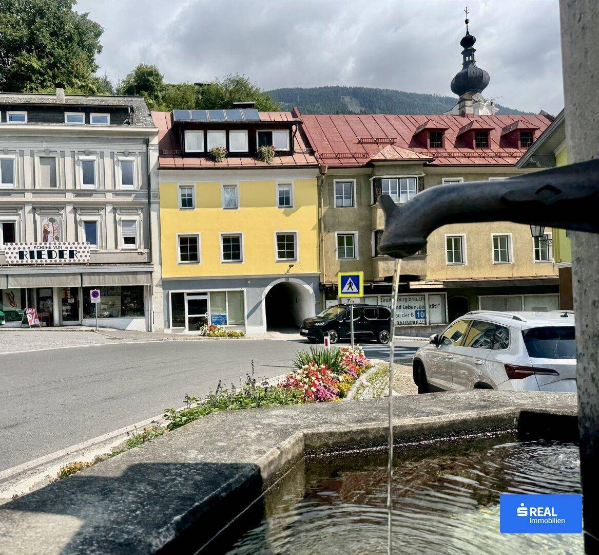 Panoramablick auf eine historische Stadtstraße mit einem Brunnen, Gebäuden und einem Kirchturm im Hintergrund.