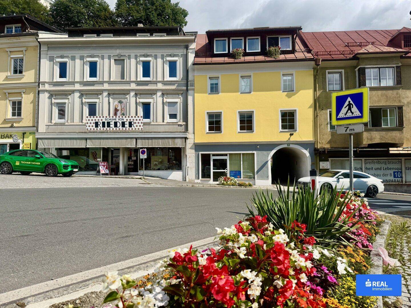 Blick auf eine belebte Straße mit farbenfrohen Gebäudefassaden und blühenden Blumenbeeten im Vordergrund.