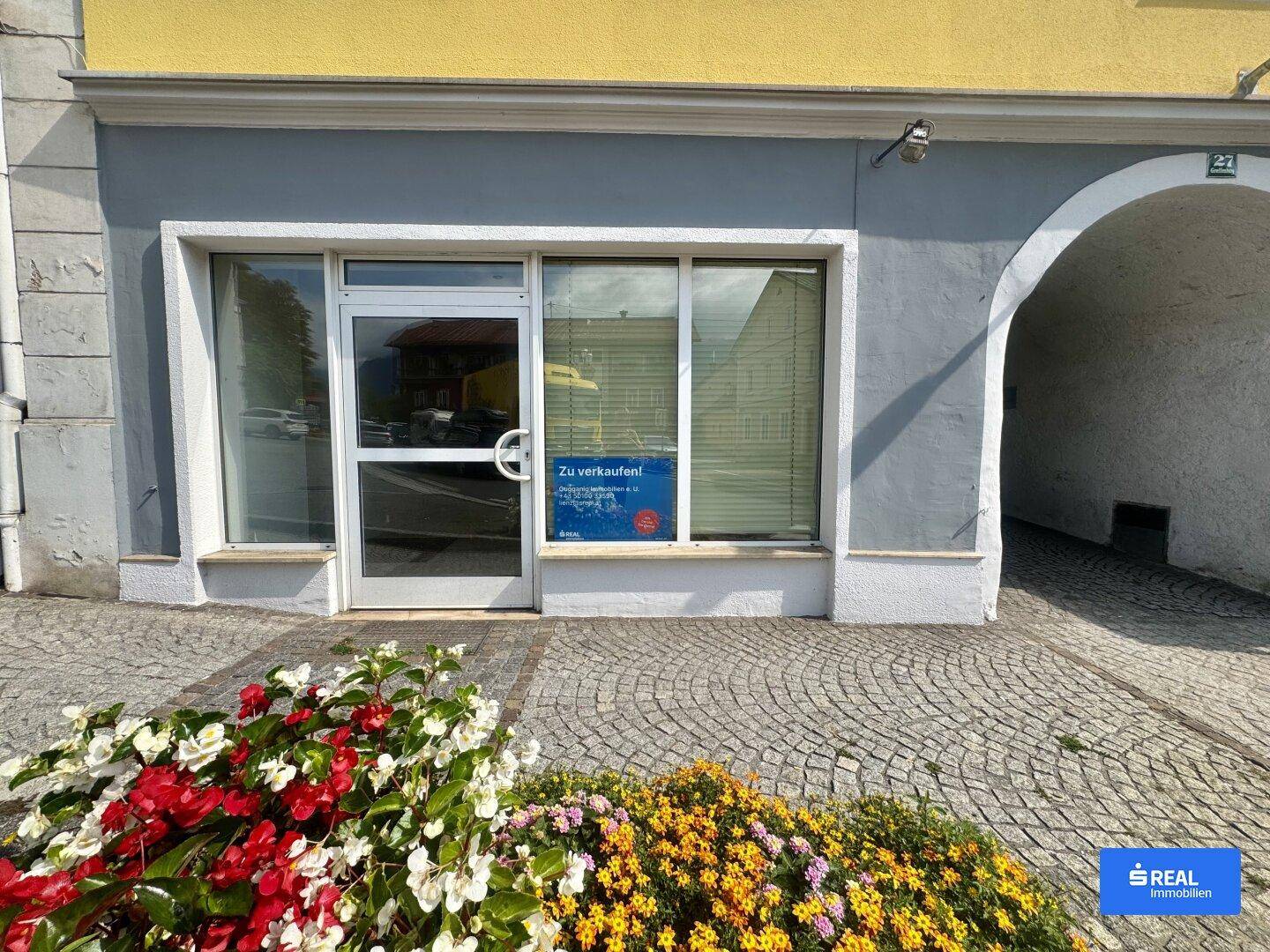 Nahaufnahme einer Ladenfront mit einem 'Zu verkaufen!'-Schild und einem angrenzenden Torbogen in einer Stadtstraße.