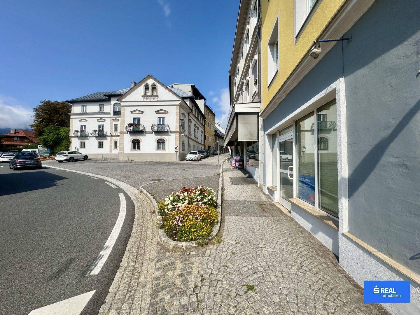 Eine gepflasterte Straßenecke mit einem Blumenbeet und traditionellen Gebäuden unter blauem Himmel.