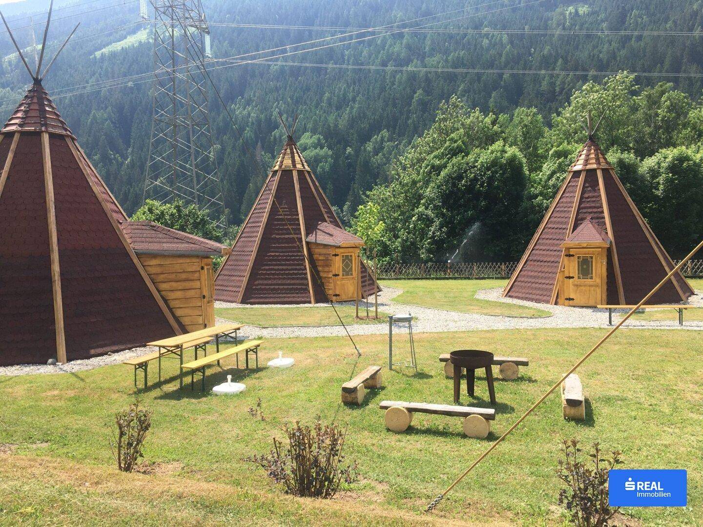 Drei Zeltstrukturen in einer grünen Landschaft mit Sitzgelegenheiten und Feuerstelle vor Bergkulisse.