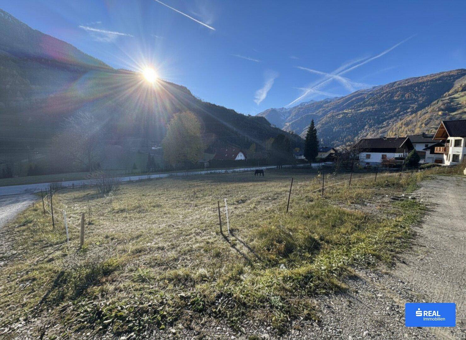 Weitläufiges, sonniges Grundstück mit Bergblick, umgeben von grünen Wiesen und einem Schotterweg.