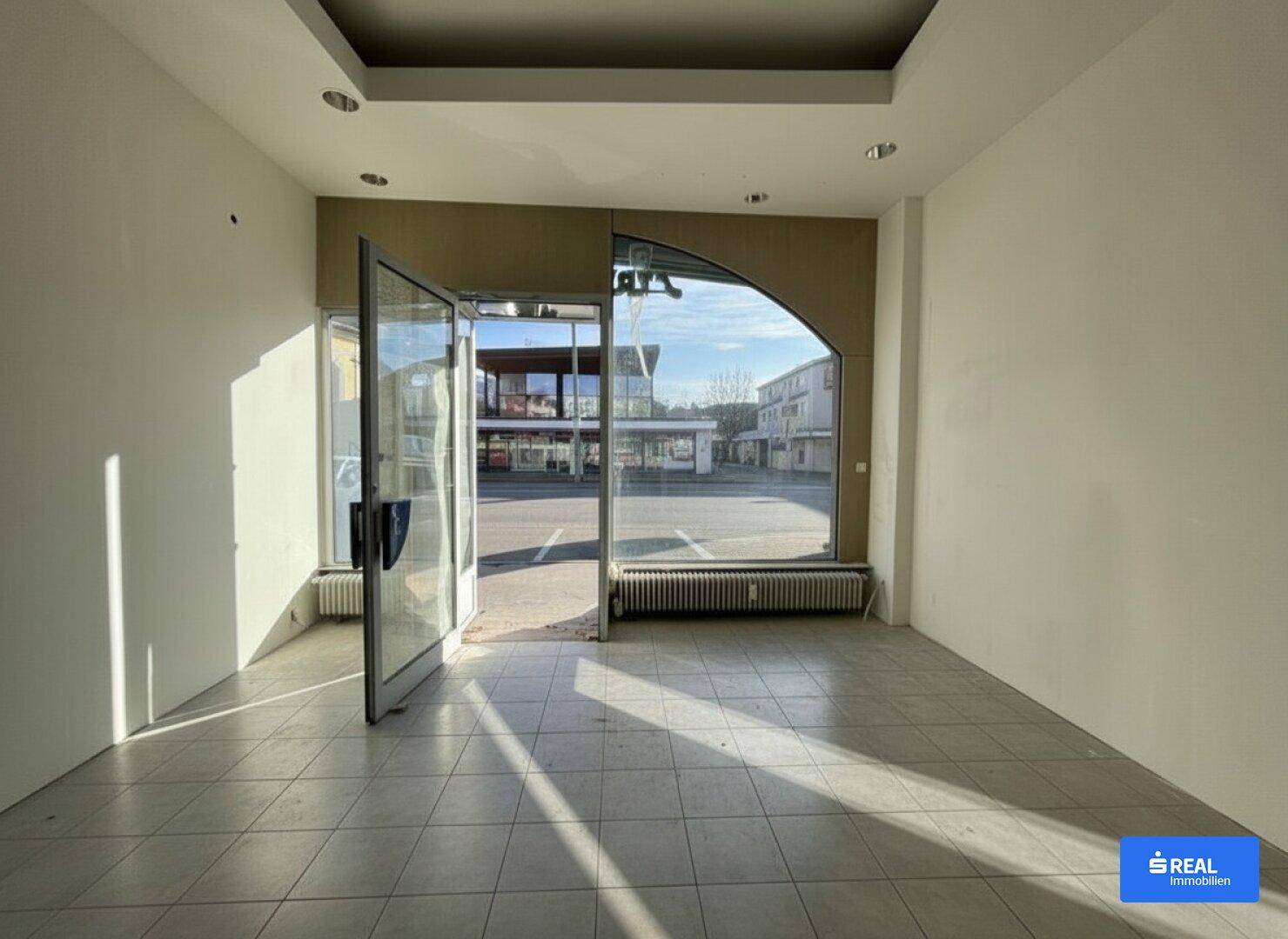 Heller Geschäftsraum im Erdgeschoss mit großen Schaufenstern und Fliesenboden, Blick zur Straße.