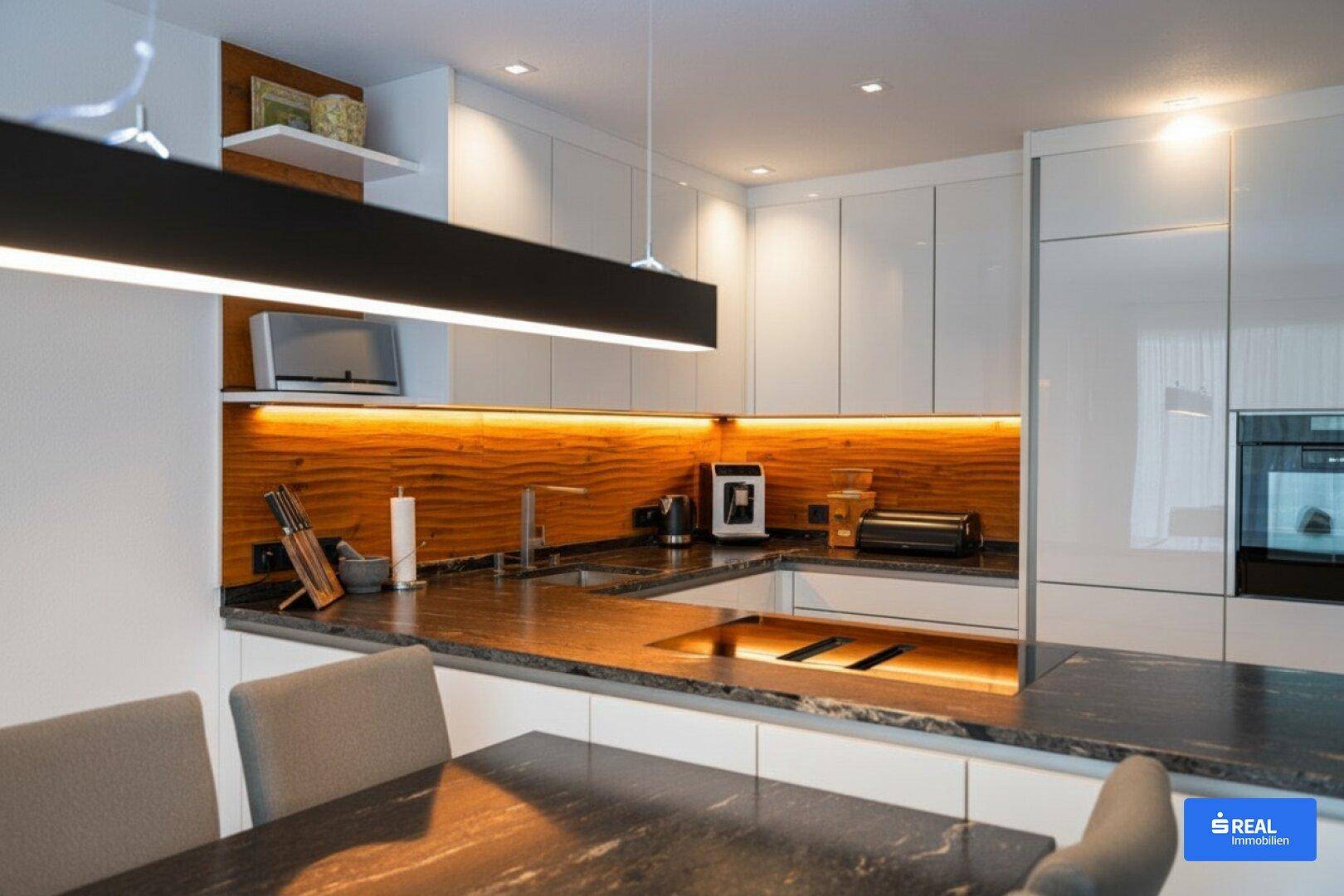 Moderne Küche mit weißen Hochglanzfronten, dunkler Arbeitsplatte und beleuchteter Holzrückwand.
