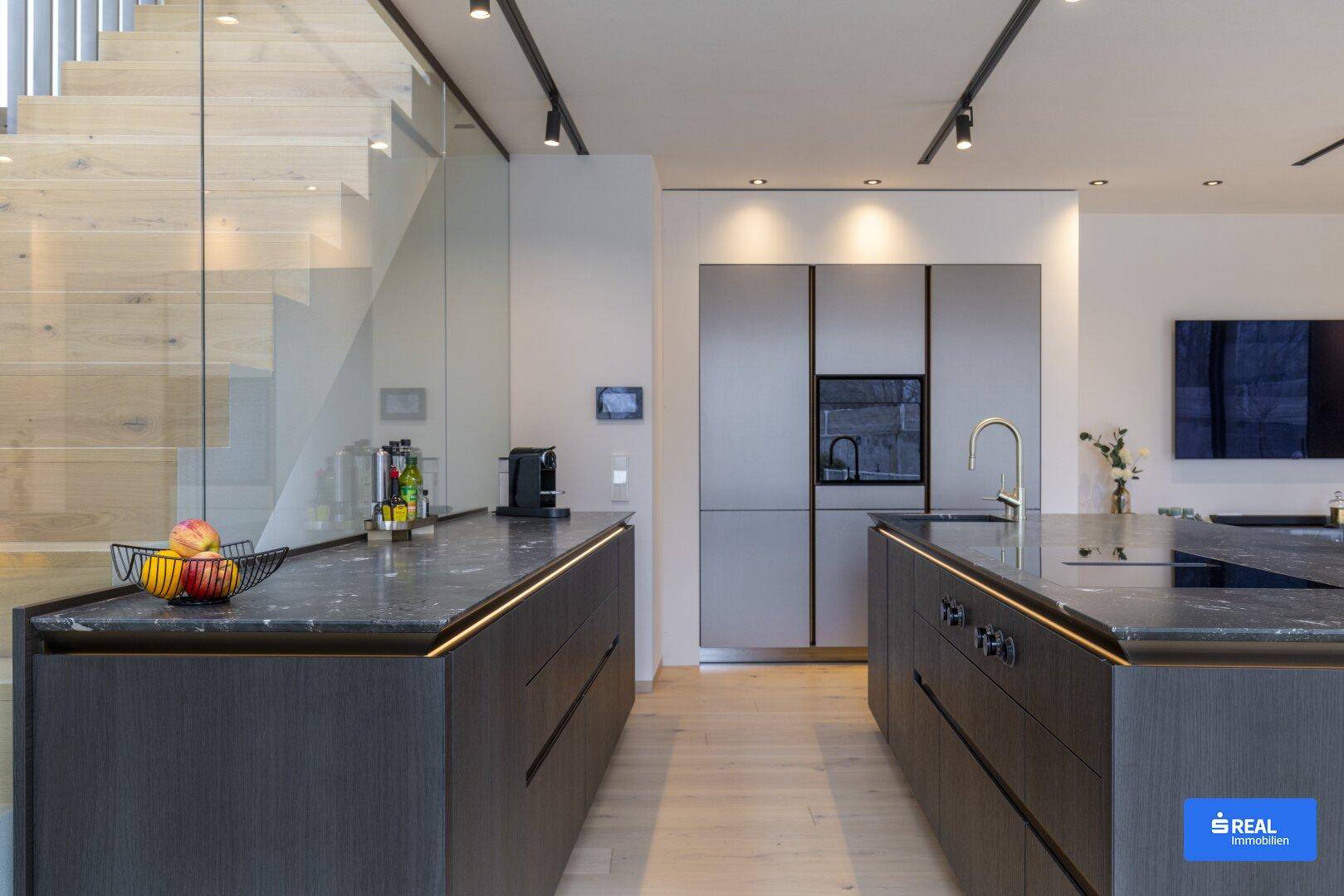 Moderne Küche mit zwei Kücheninseln, dunklen Arbeitsplatten und integrierten Geräten unter einer eleganten Treppe.