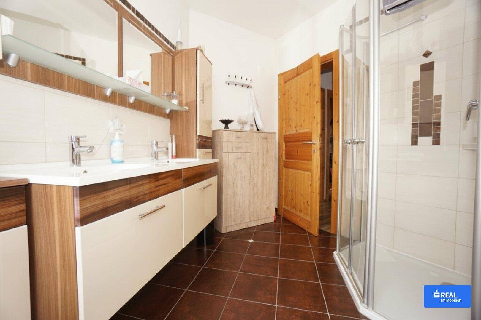 Badezimmer mit Doppelwaschbecken, Duschkabine und braunen Fliesen.